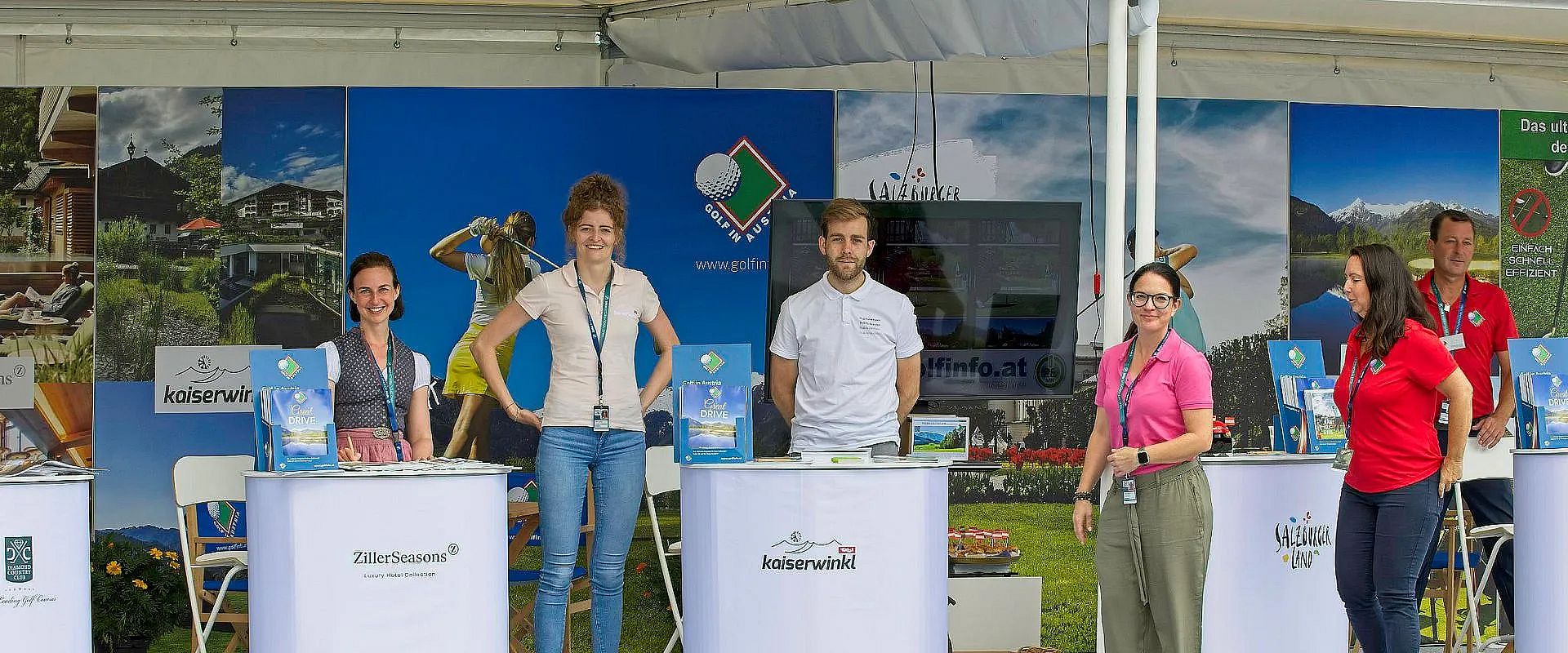 Vier Personen stehen an Messeständen mit Golf-Thema. Im Hintergrund sind Golfposter und ein Bildschirm zu sehen. Sie tragen Badge-Karten um den Hals.