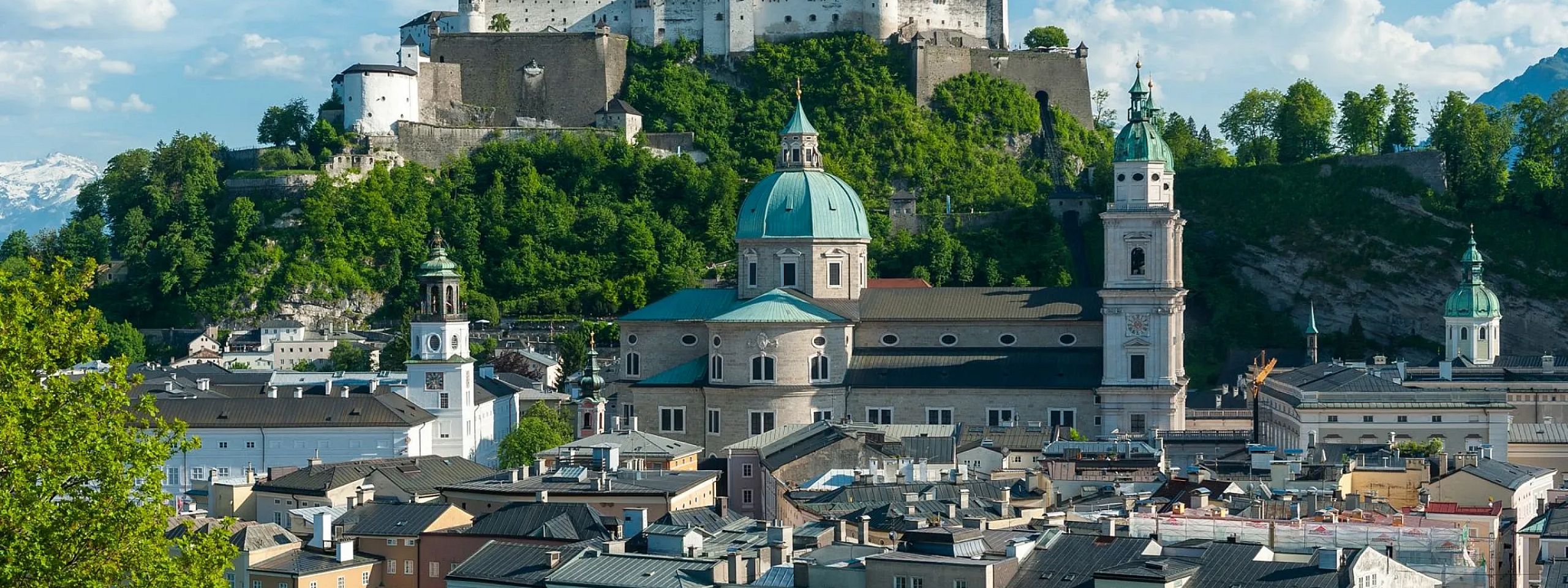 Panorama von Salzburg mit der Festung Hohensalzburg im Hintergrund und der barocken Architektur der Stadt im Vordergrund, umgeben von grünen Hügeln.