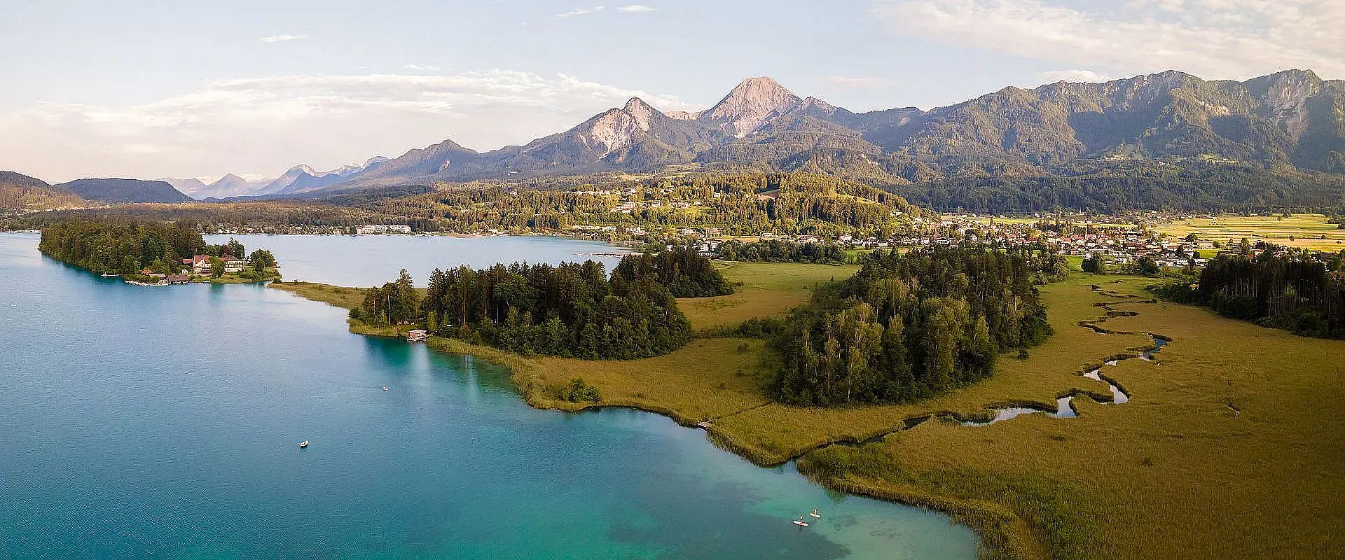Panoramablick auf einen malerischen See, umgeben von üppig grüner Landschaft und majestätischen Bergen im Hintergrund, unter einem klaren Himmel.