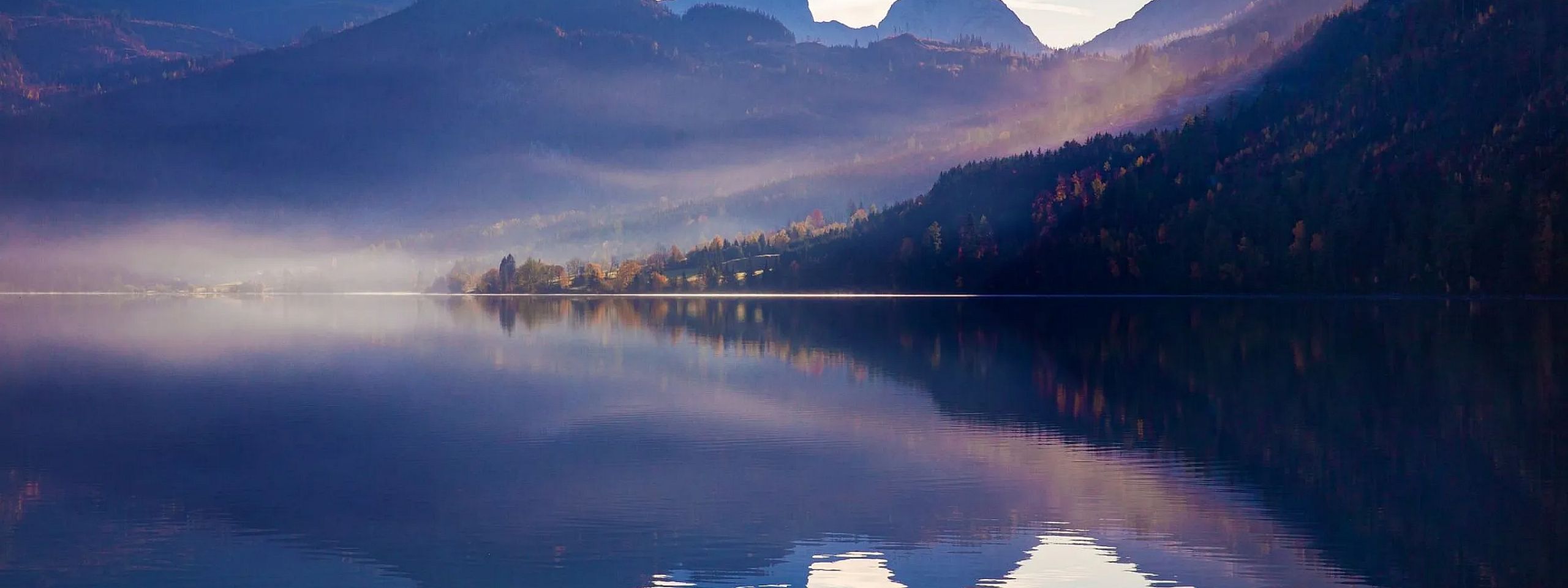 Malerische Berglandschaft mit Nebel über einem ruhigen See, umgeben von herbstlichen Bäumen. Sanfte Morgen- oder Abenddämmerung beleuchtet die Szene.