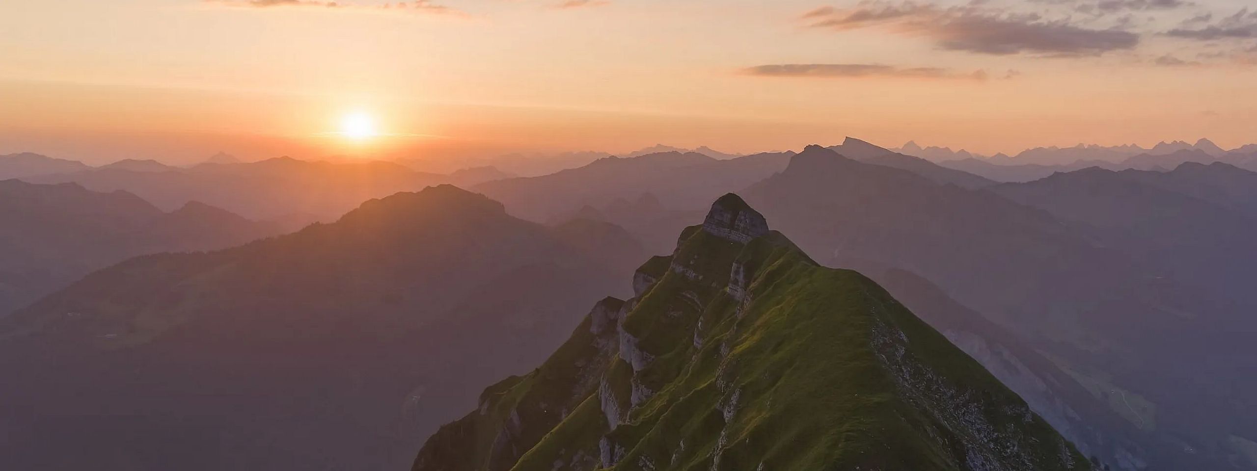 Ein malerischer Sonnenuntergang über einer Bergkette mit grünen Hängen. Die Sonne steht tief am Himmel und taucht die Landschaft in ein warmes, goldenes Licht.