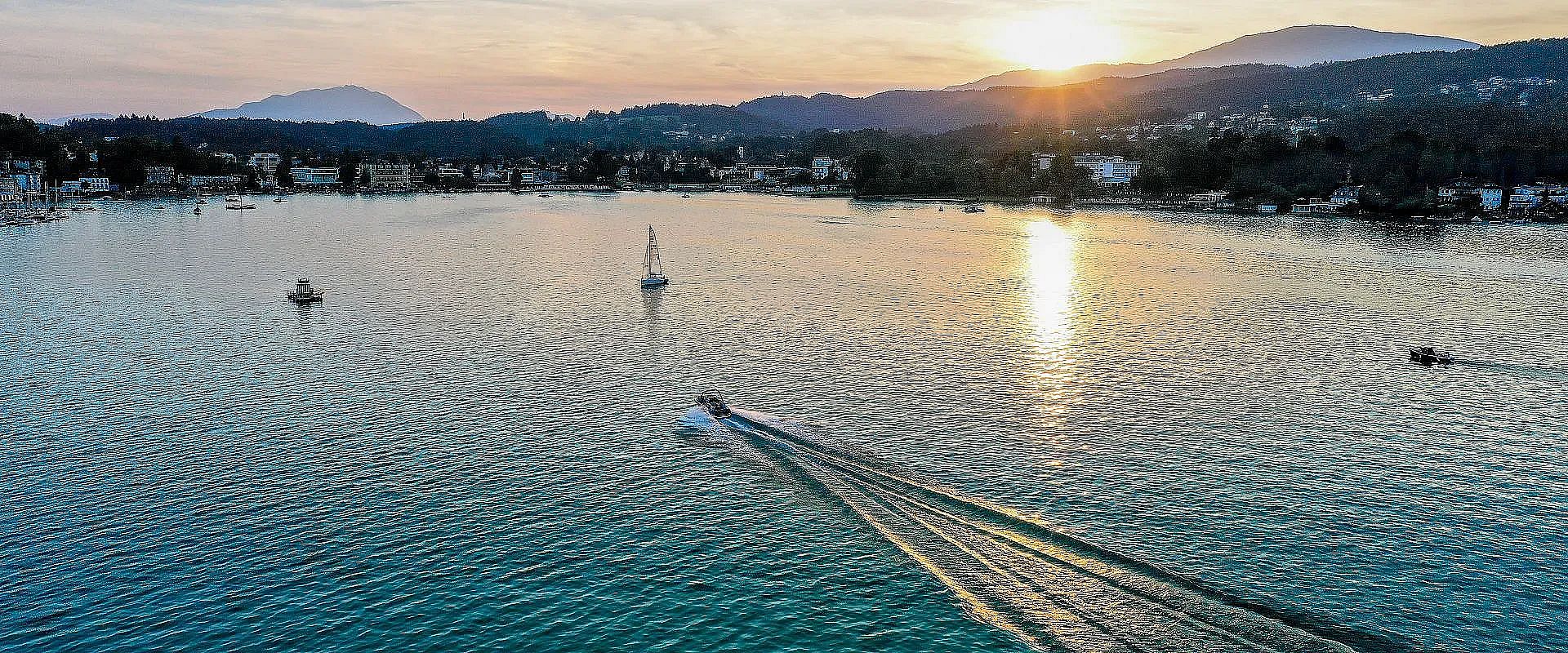 Abendstimmung am See mit einem Boot, das Spuren im Wasser zieht. Die Sonne geht hinter Hügeln unter, während im Hintergrund Berge und Uferhäuser zu sehen sind.