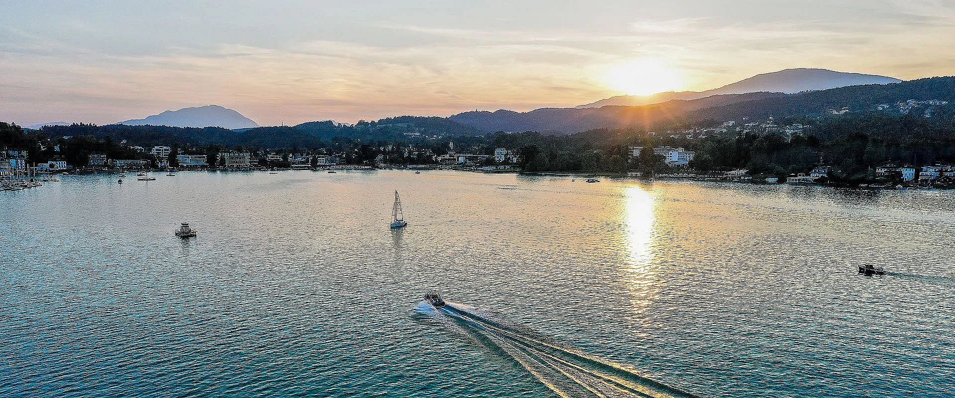 Ein ruhiger See bei Sonnenuntergang mit einem Boot, das auf dem Wasser eine Spur hinterlässt. Im Hintergrund sind bewaldete Hügel und ein heller Himmel.