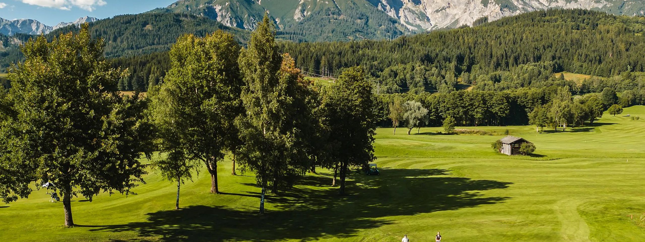 Ein weitläufiger Golfplatz mit grünen Fairways und Bunkern, umgeben von Bäumen. Im Hintergrund erheben sich majestätische Berge unter einem blauen Himmel.