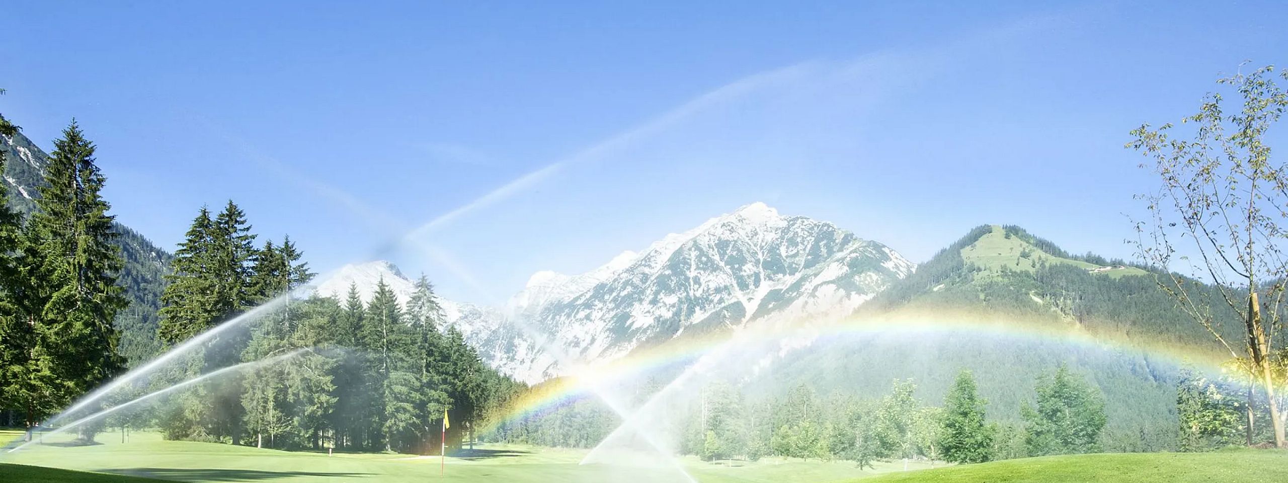 Ein weitläufiges grünes Feld mit Rasensprinklern, die Wasser in die Luft sprühen. Im Hintergrund sind hohe Berge und ein klarer blauer Himmel zu sehen.