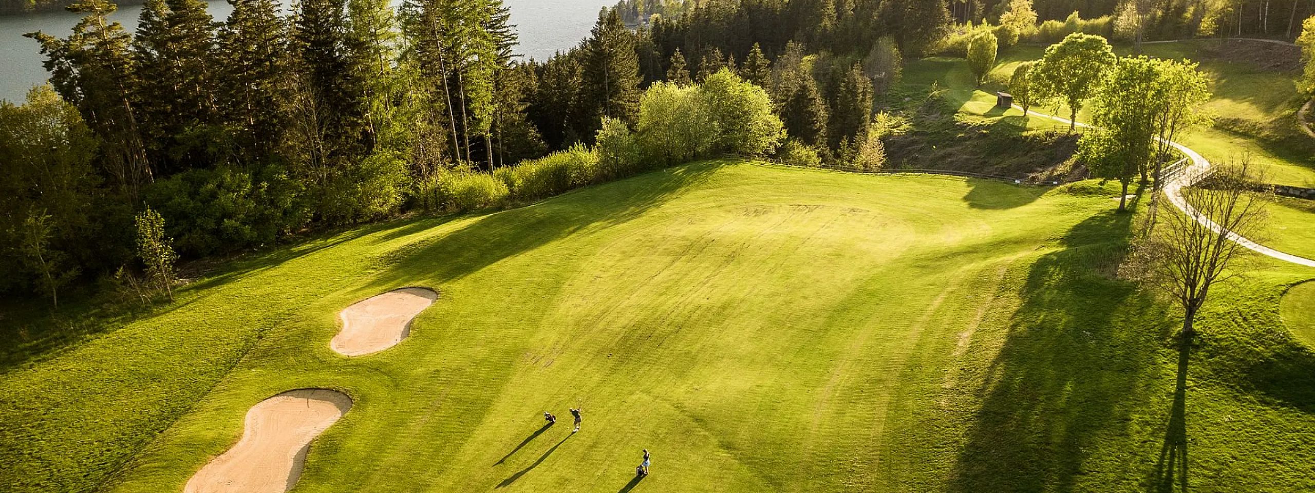 Grüner Golfplatz mit zwei Sandbunkern, umgeben von dichtem Wald und in der Ferne ein See. Zwei Personen stehen auf dem Rasen unter klarem Himmel.