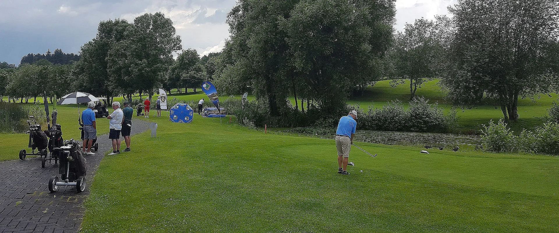 Ein Mann in hellem Polohemd und Shorts steht auf einem Golfplatz bereit zum Abschlag. Eine kleine Gruppe von Zuschauern beobachtet ihn neben grünen Bäumen.