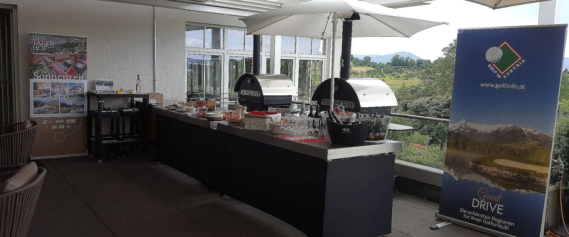 Moderne Terrasse mit zwei Pizzaöfen auf einem Tisch. Rundherum stehen Getränke und Zutaten. Im Hintergrund sind Fenster und eine grüne Landschaft zu sehen.