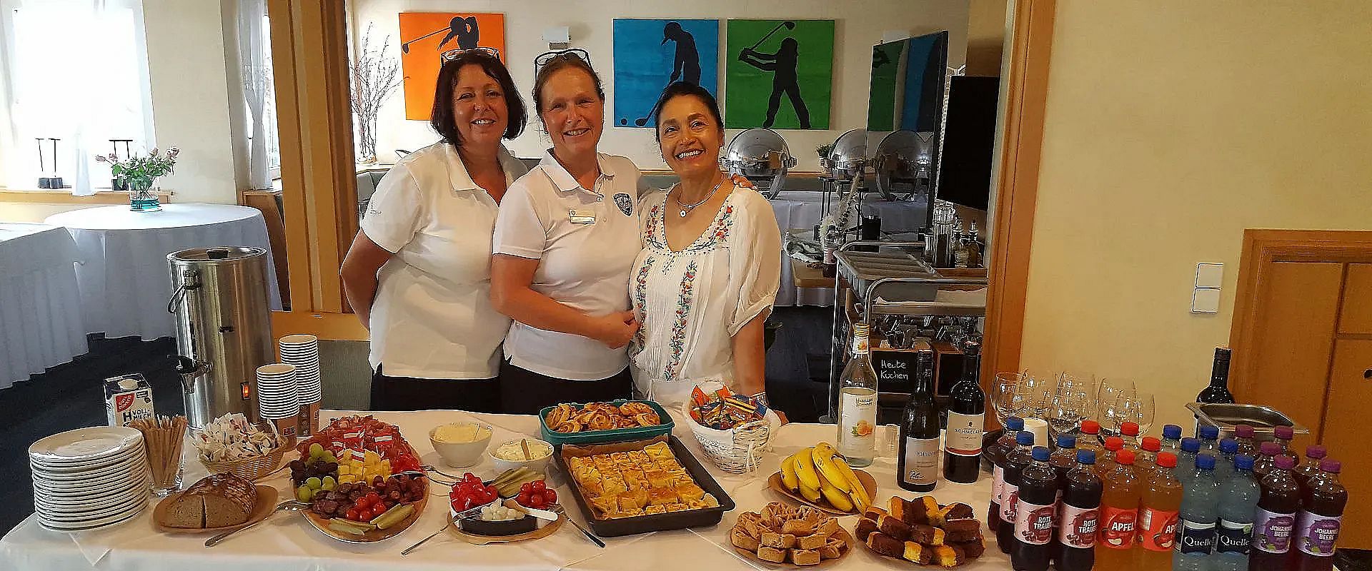 Drei Frauen stehen hinter einem Tisch mit einem Buffet aus Obst, Gebäck und Getränken in einem hellen Raum mit bunten Kunstwerken an den Wänden.