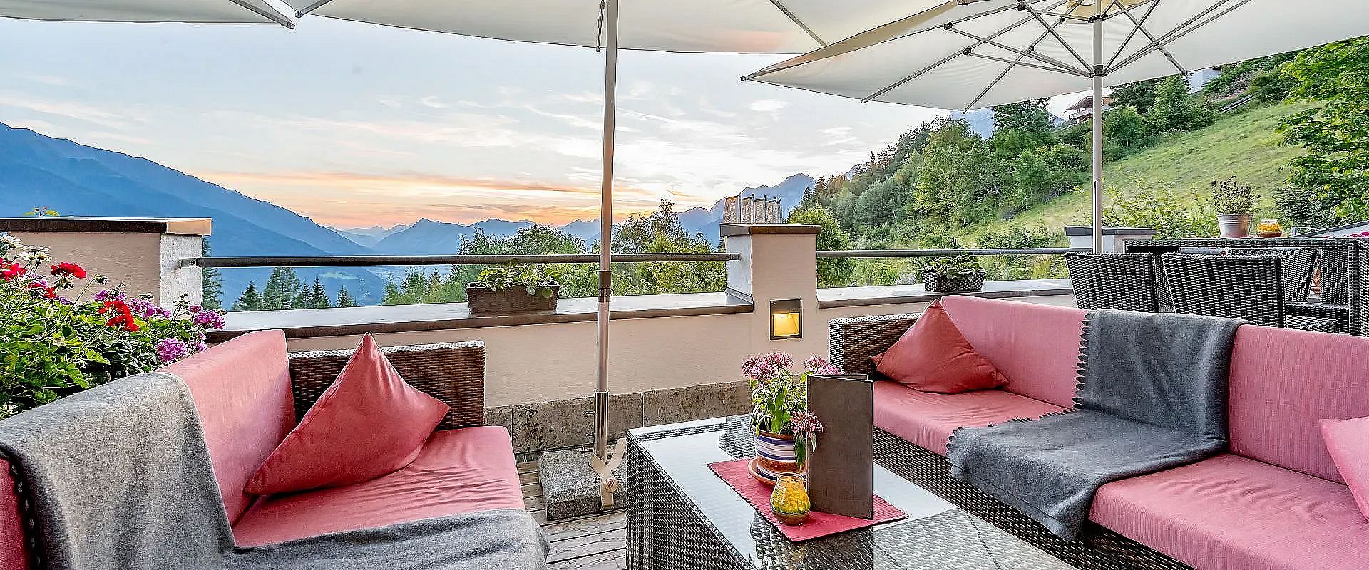 Gemütliche Terrasse mit roten Polsterbänken und Glas-Couchtisch, umgeben von Bergen. Der Sonnenuntergang taucht den Himmel in warme Farben. Sonnenschirme spenden Schatten.