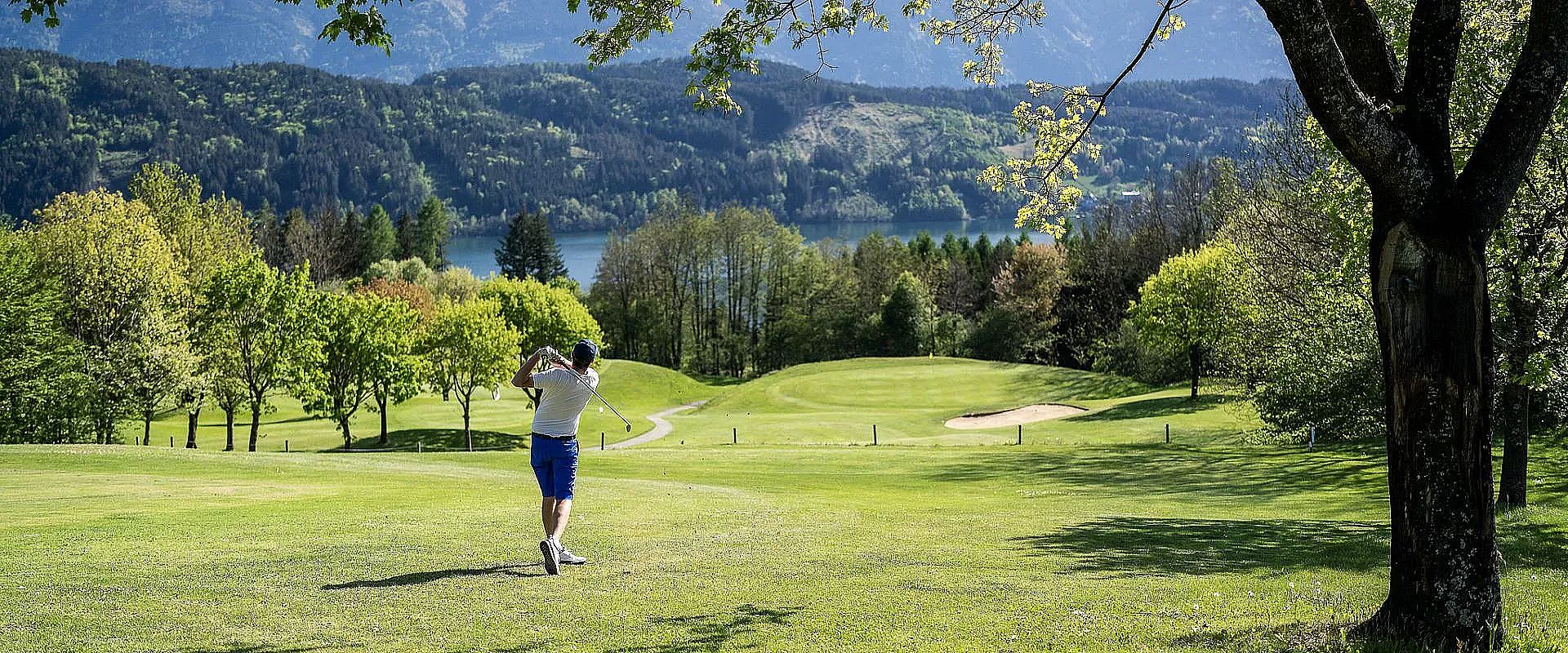 Ein Golfer schlägt einen Ball auf einem gepflegten Golfplatz mit grünen Bäumen und Blick auf einen See und Berge im Hintergrund an einem sonnigen Tag.