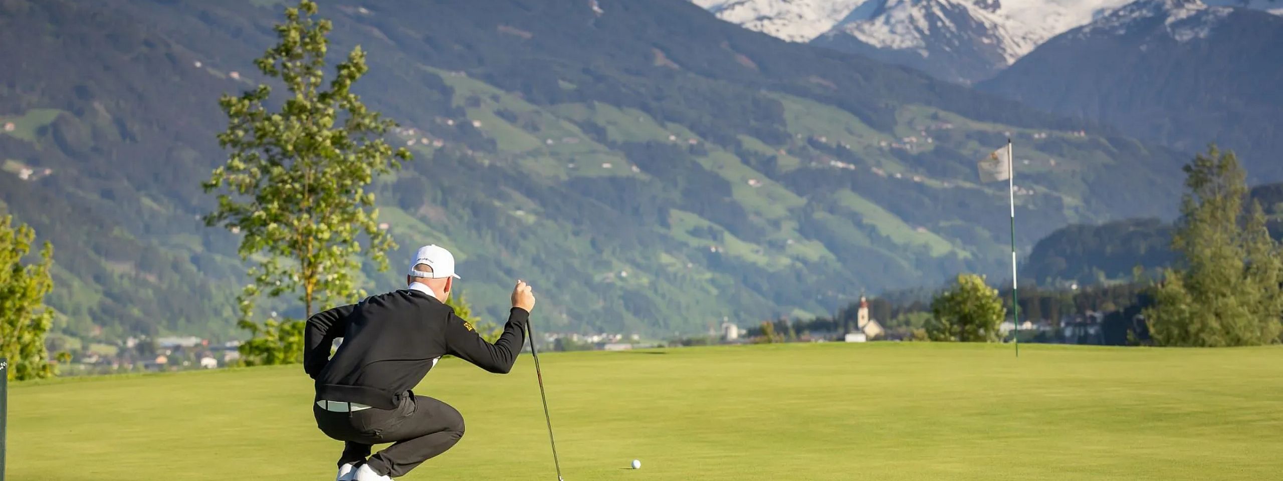 Ein Golfer in schwarzem Outfit und weißer Kappe kniet auf einem Golfplatz und zielt auf einen Ball. Im Hintergrund sind Berge mit schneebedeckten Gipfeln sichtbar.
