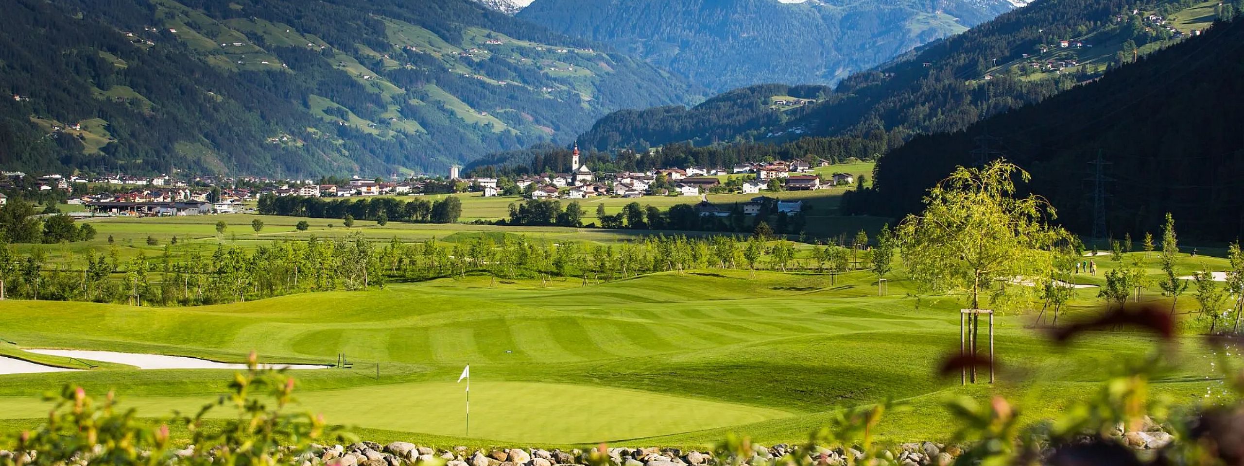 Blick auf einen Golfplatz im Tal mit gemähten Fairways, umgeben von grünen Hügeln und schneebedeckten Bergen im Hintergrund, im Vordergrund ein kleiner Teich und Blumen.