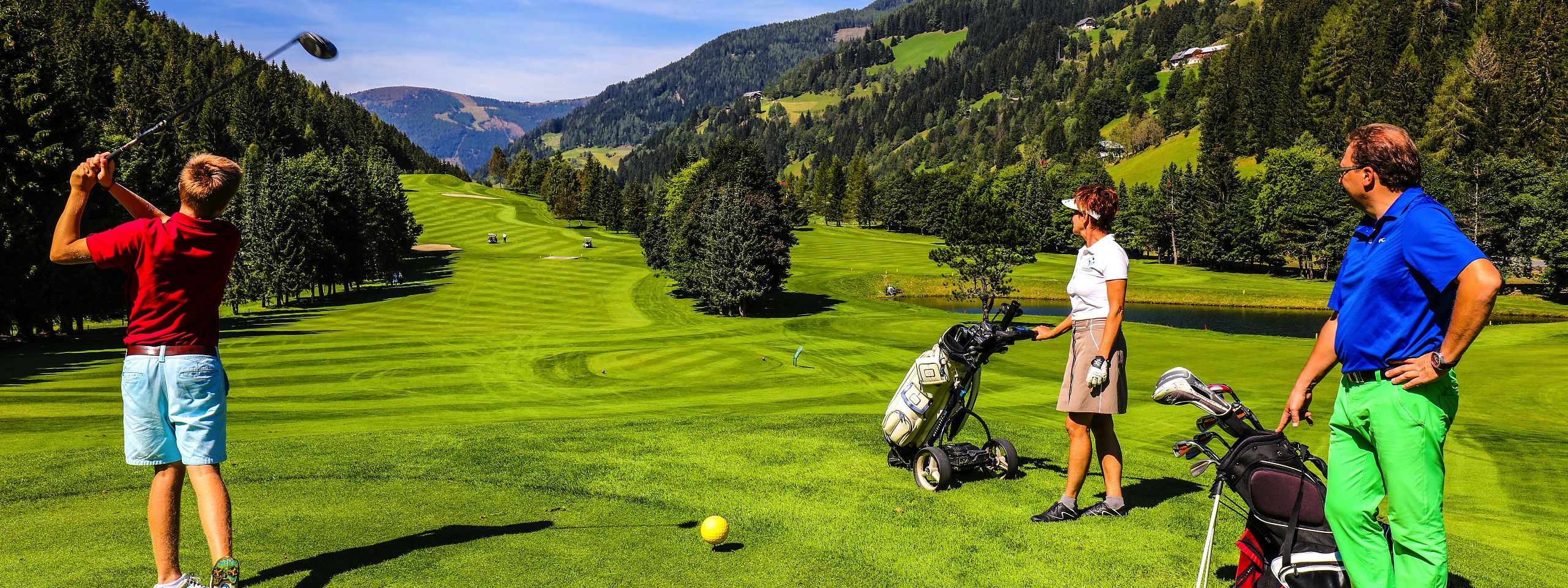 Drei Menschen spielen Golf auf einem grünen Platz in einer malerischen Berglandschaft. Eine Person schwingt den Schläger, während die anderen zuschauen.