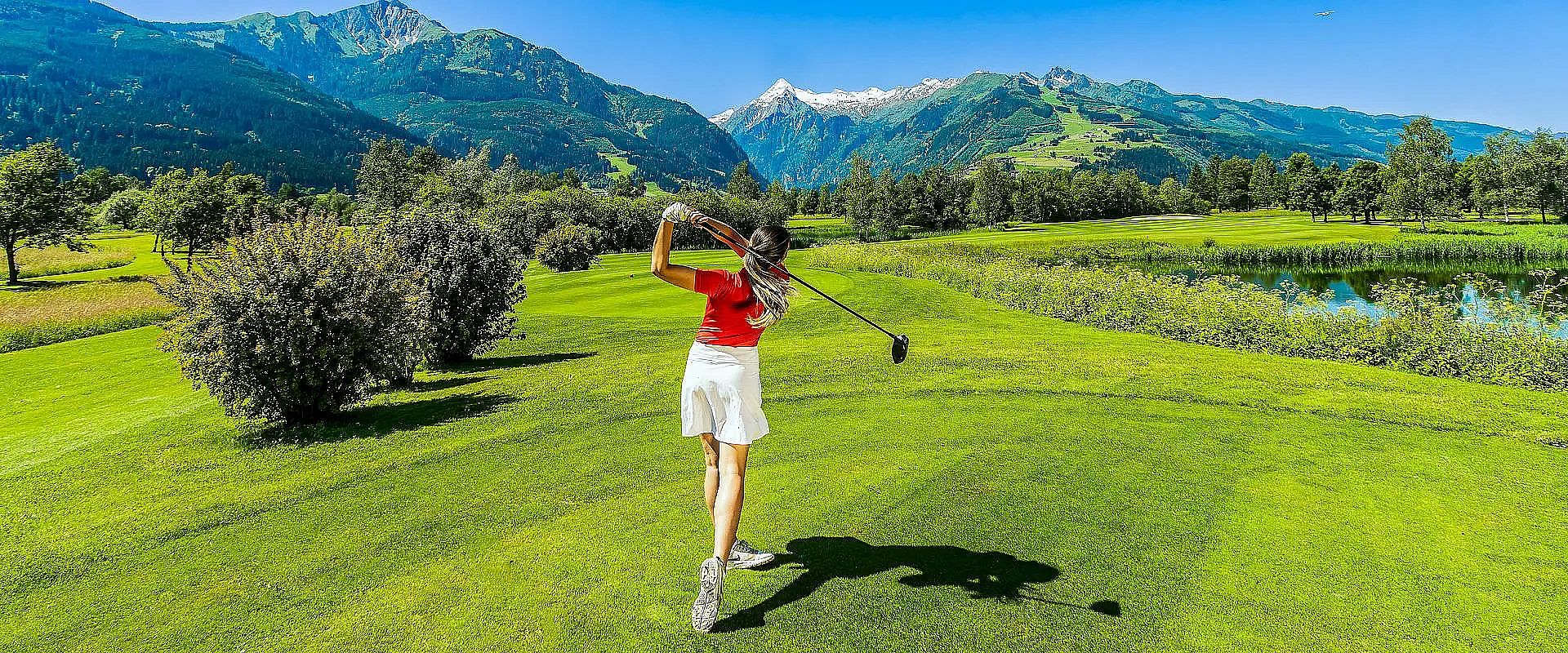 Eine Frau in sportlicher Kleidung schlägt einen Golfball auf einem grünen Fairway. Im Hintergrund sind Berge und ein klarer blauer Himmel zu sehen.