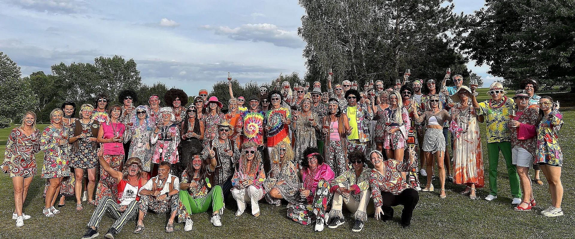 Eine große Gruppe von Menschen in bunter Hippie-Kleidung posiert fröhlich auf einer Wiese. Viele tragen auffällige Muster, Sonnenbrillen und Kostüme.
