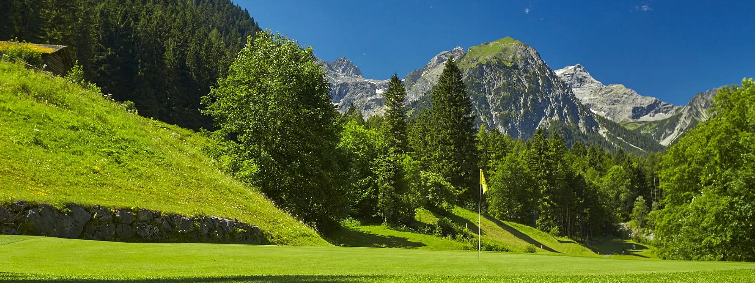 Grüne Wiese mit Fahne auf einem Golfplatz, umgeben von hohen Bäumen und majestätischen Bergen im Hintergrund unter einem klaren blauen Himmel.