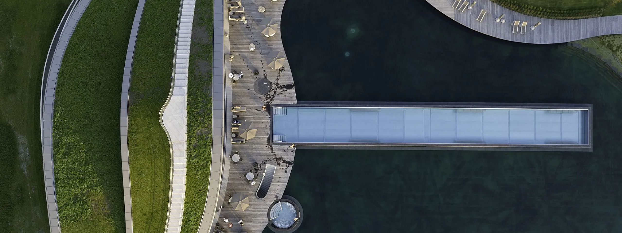 Luftaufnahme eines modernen Parks mit geschwungenen Wegen, einer zentralen Brücke über Wasser und umgeben von Grasflächen und Sitzbereichen mit Sonnenschirmen.