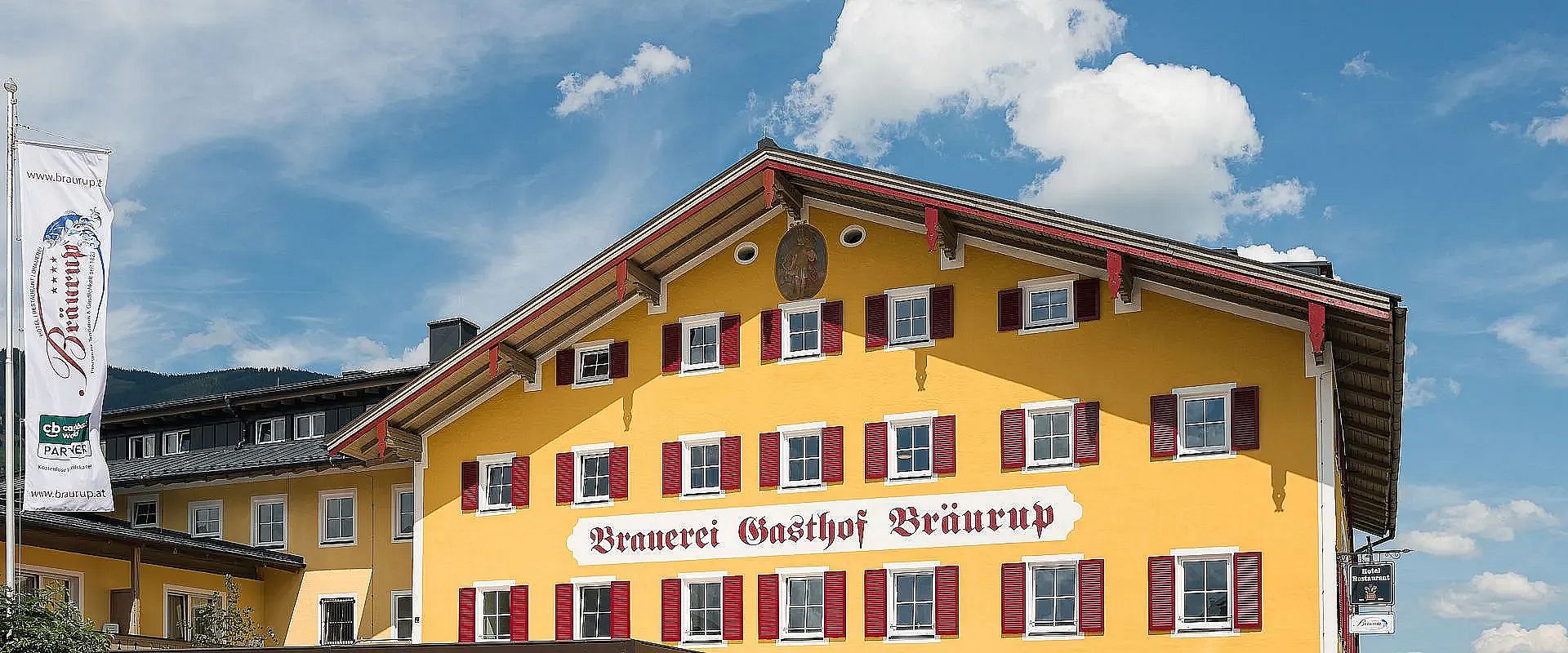 Ein traditionelles bayerisches Gebäude mit gelber Fassade und roten Fensterläden, beschriftet mit "Brauerei Gasthof Bräurup", umgeben von blauem Himmel.