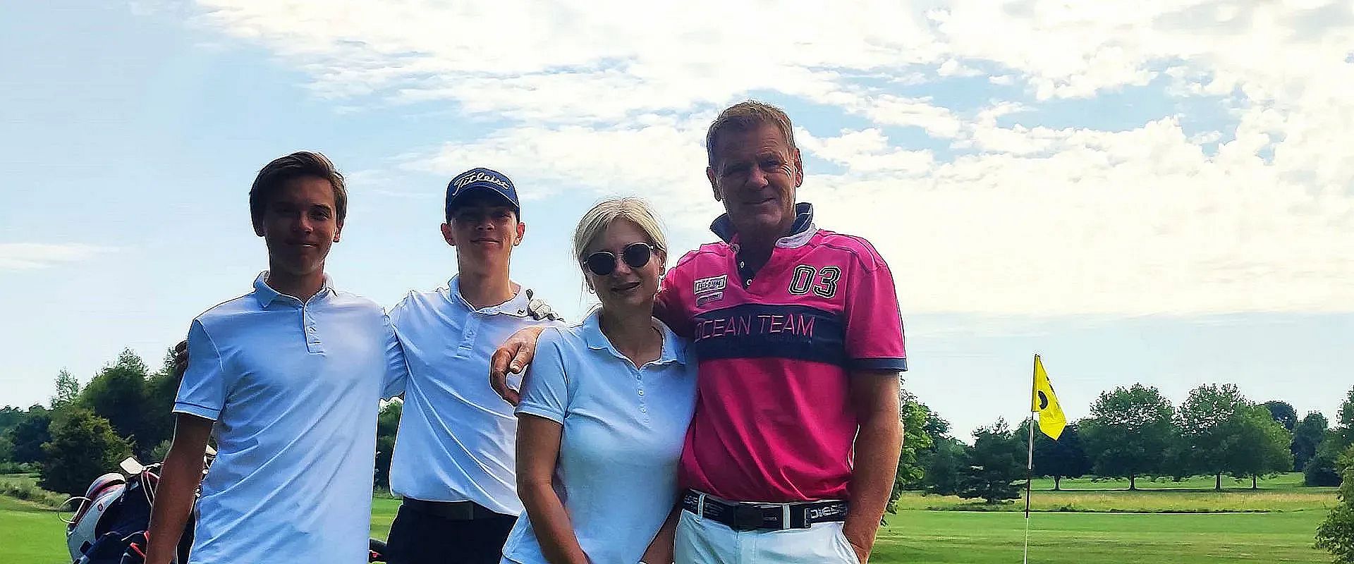 Vier Personen auf einem Golfplatz posieren lächelnd für ein Foto. Sie tragen sportliche Kleidung, und im Hintergrund sind Golffahnen und ein klarer Himmel zu sehen.