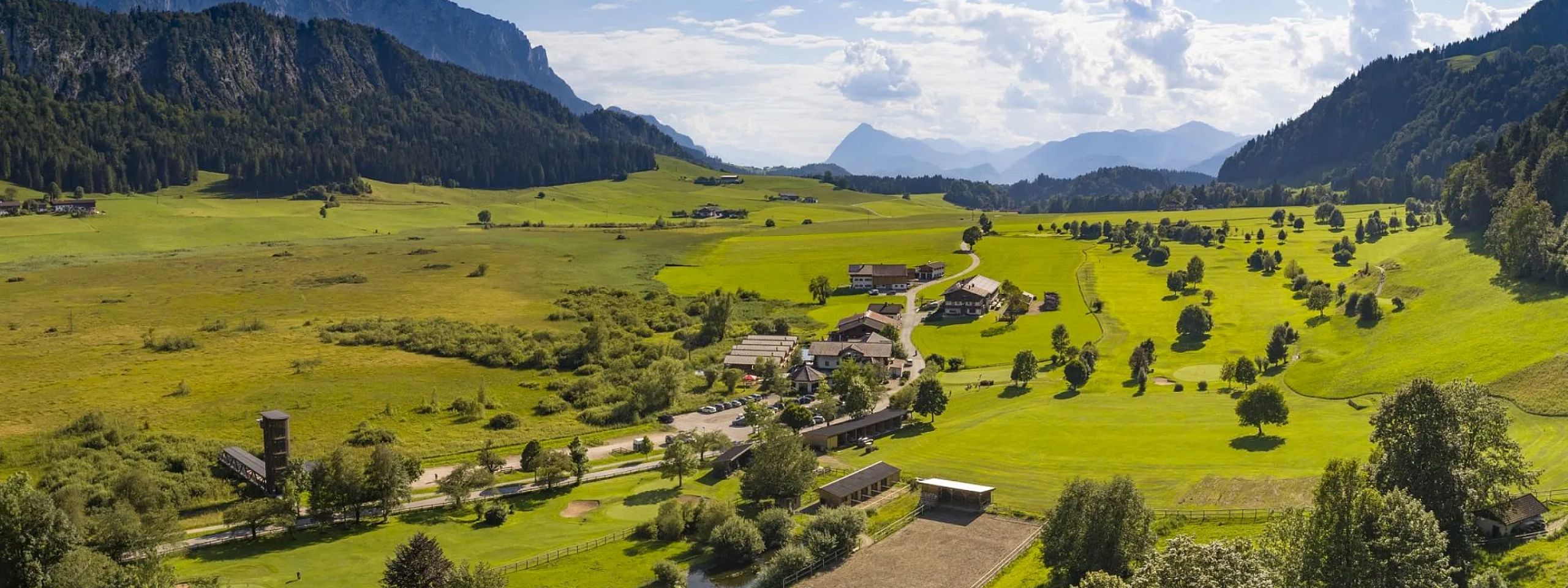 Panorama einer grünen Landschaft in den Bergen, mit einem Dorf im Tal. Im Hintergrund sind bewaldete Hänge und Gipfel unter einem bewölkten Himmel zu sehen.
