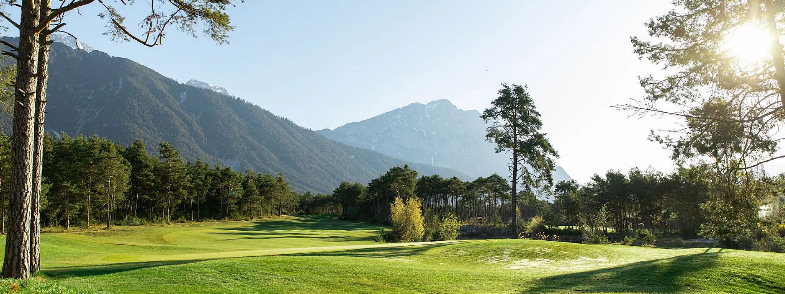 Eine malerische Landschaft mit einem grünen Golfplatz im Vordergrund, umgeben von hohen Bäumen und majestätischen Bergen im Hintergrund unter klarem Himmel.