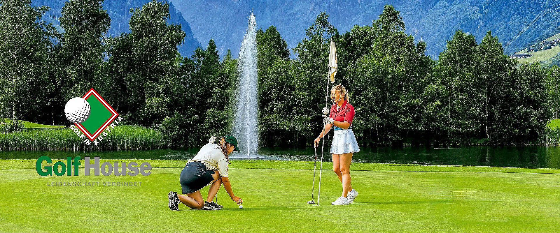 Zwei Personen auf einem Golfplatz, eine kniet, um einen Ball zu platzieren, während die andere den Flaggenstock hält. Im Hintergrund sind Bäume und ein Wasserfall.