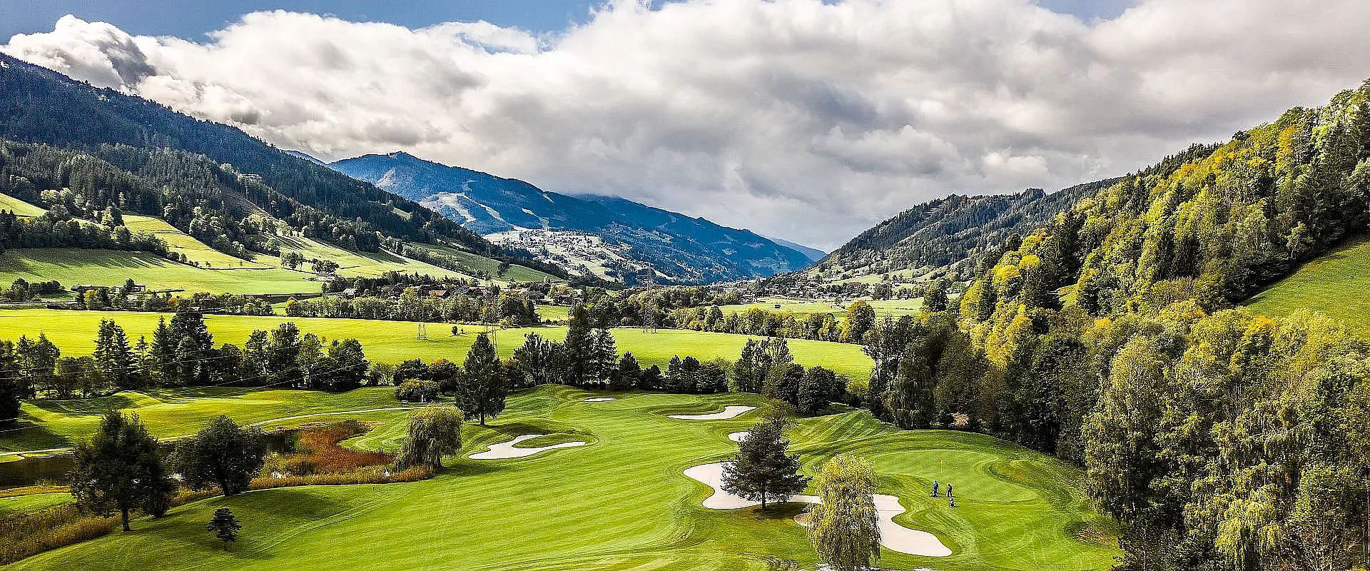 Idyllische Golfplatzlandschaft umgeben von Hügeln und bewaldeten Bergen. Wolken ziehen über den blauen Himmel, während das grüne Gras lebendig leuchtet.