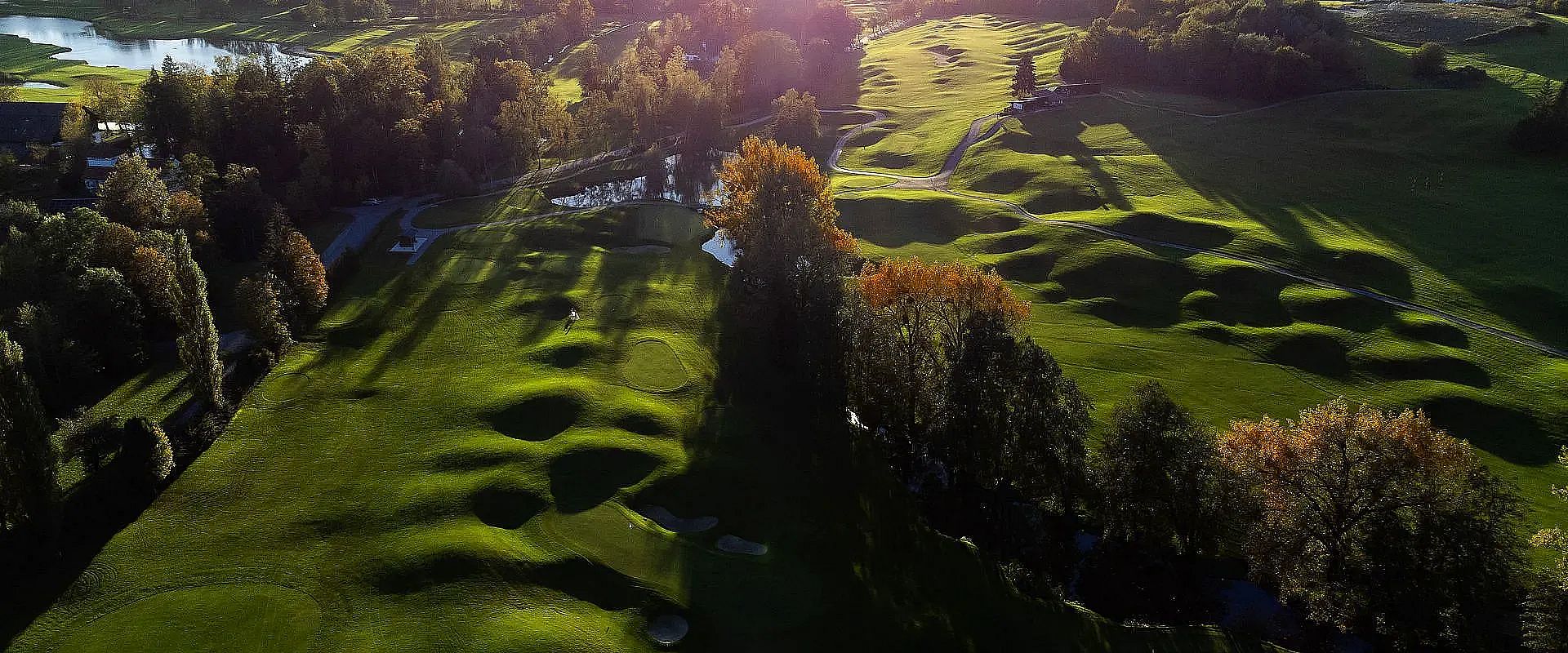 Luftaufnahme eines grünen Golfplatzes bei Sonnenuntergang, umgeben von Bäumen. Das Licht erzeugt lange Schatten auf den welligen Fairways und Greens.