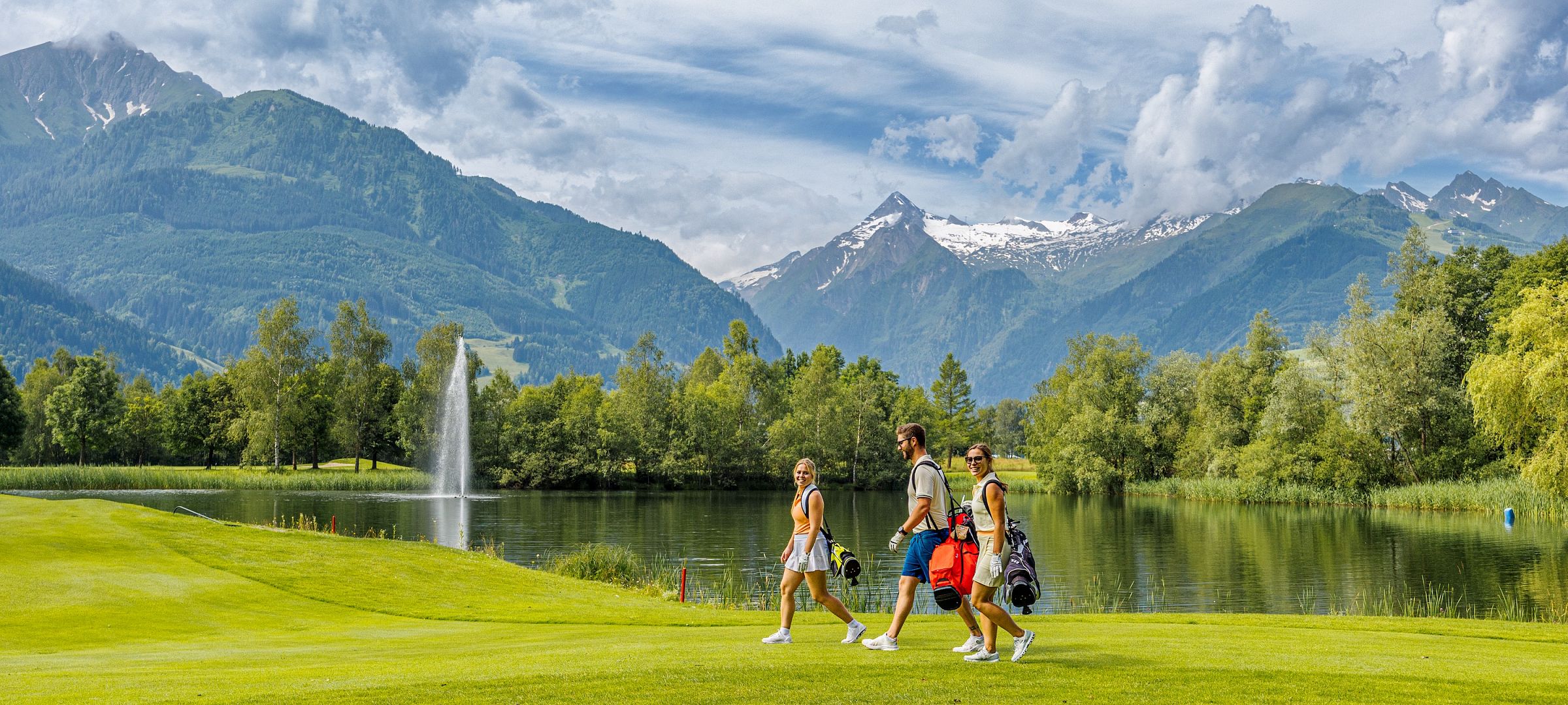 Drei Golfer gehen über einen grünen Golfplatz mit Bergen im Hintergrund. Ein See mit einem Springbrunnen ist sichtbar, und der Himmel ist teils bewölkt.