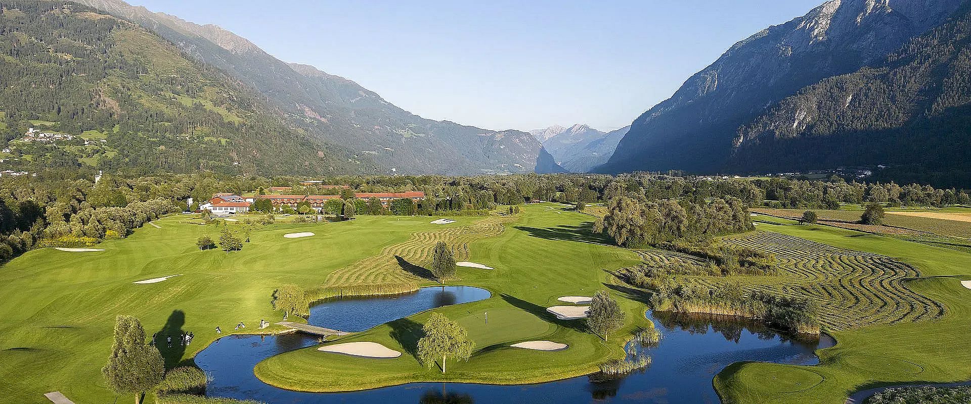 Panoramablick auf einen grünen Golfplatz inmitten von Bergen. Der Platz erstreckt sich zwischen sanften Hügeln und Wasserhindernissen, umgeben von Alpenlandschaft.