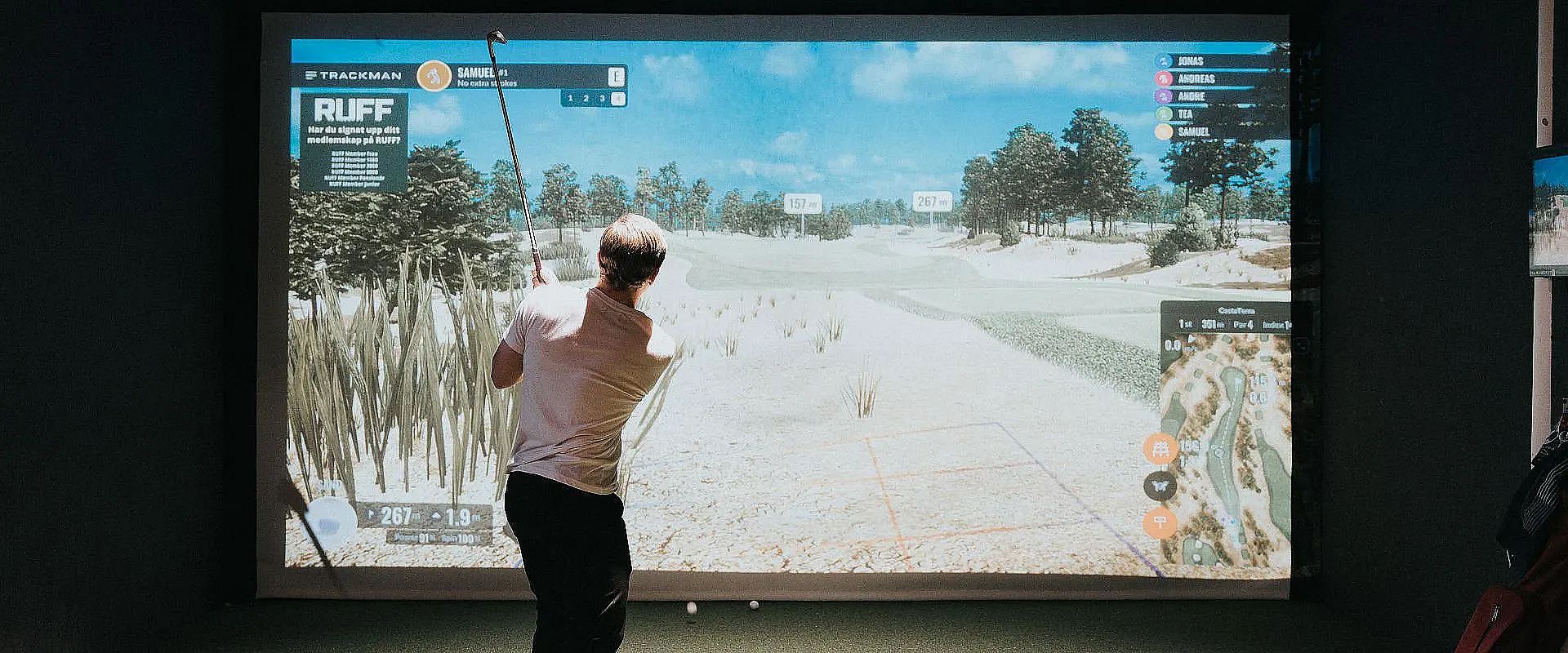 Ein Mann spielt Golf in einem Indoor-Simulatorraum. Der Bildschirm zeigt eine virtuelle Golfbahn mit Bäumen und Sandbunkern. Er trägt ein weißes Hemd.