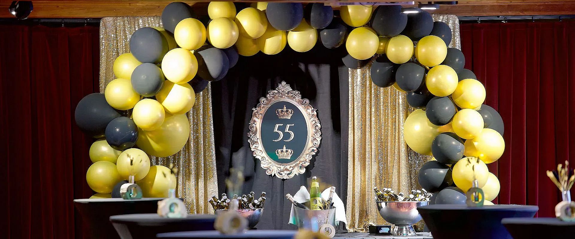 Festlich dekorierter Raum mit goldenen und schwarzen Luftballons, goldenen Vorhängen und einem Schild mit der Zahl 55. Elegantes Ambiente für eine Feier.