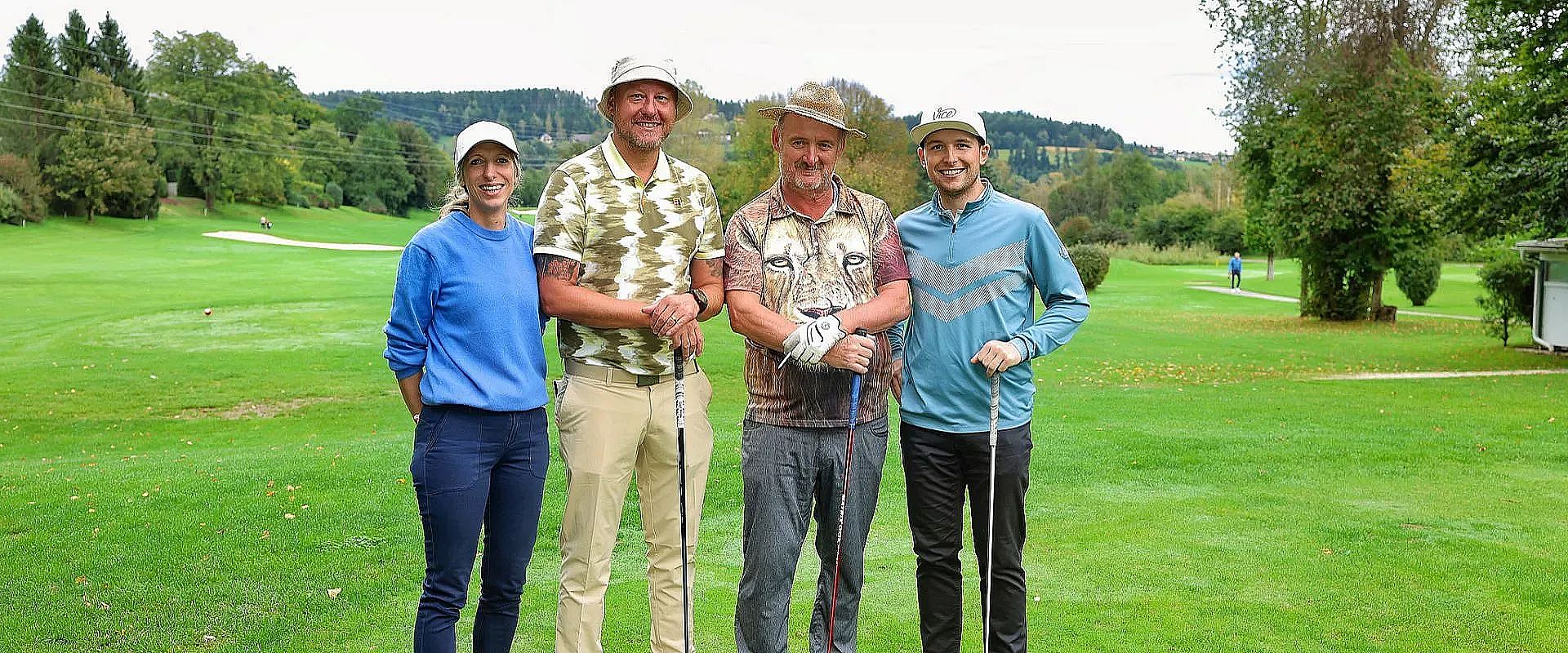 Vier Personen stehen auf einem Golfplatz. Es sind zwei Männer und zwei Frauen in sportlicher Kleidung. Sie halten Golfschläger und lächeln in die Kamera.
