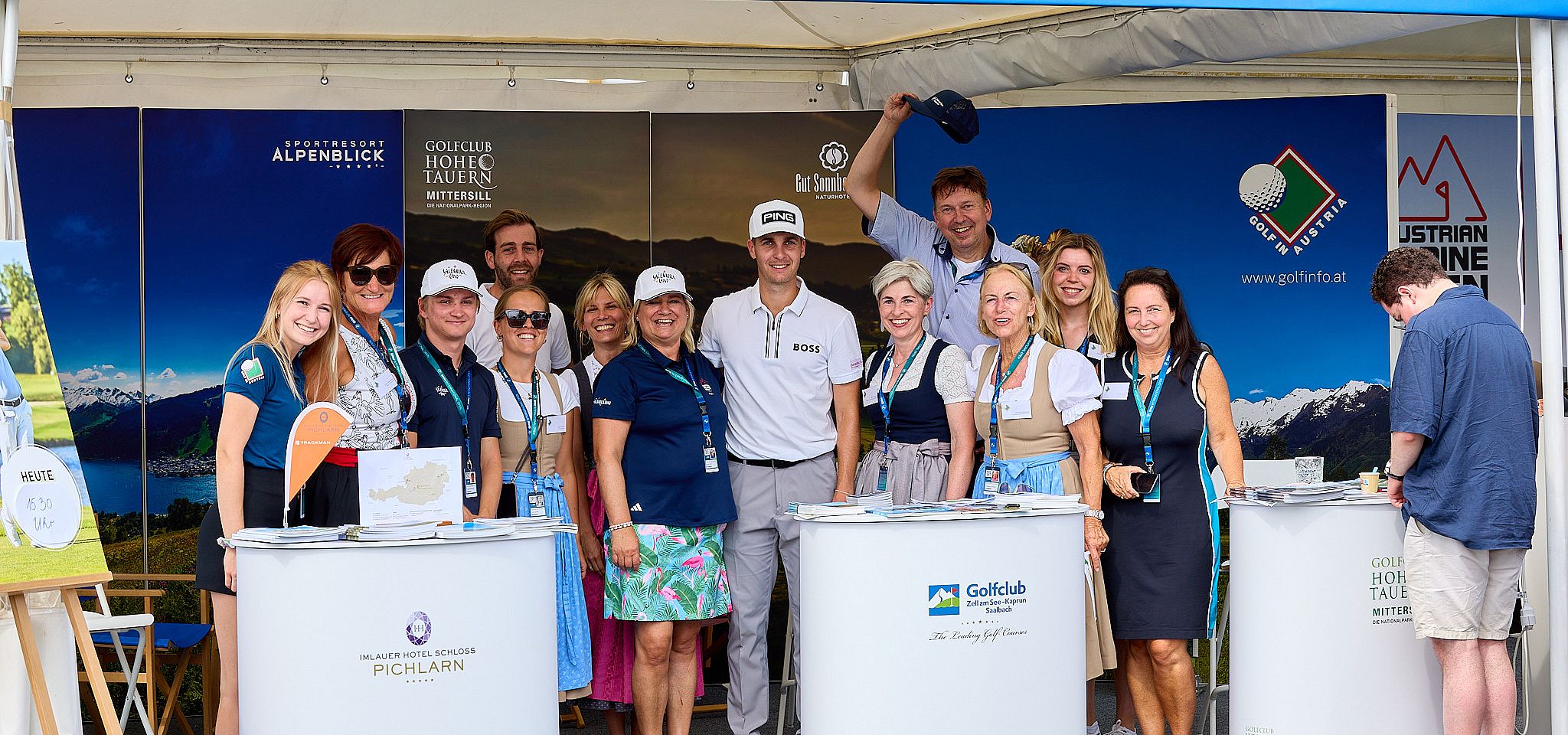 Eine Gruppe von Menschen in traditioneller Kleidung lächelt in die Kamera. Ein Mann in Golfkleidung steht zentral, umgeben von anderen Personen.