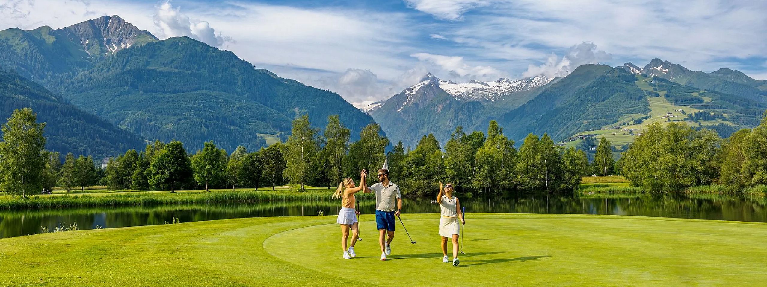 Drei Personen spielen Golf auf einem saftigen grünen Platz vor beeindruckender Bergkulisse. Der Himmel ist teils bewölkt, und die Sonne scheint.