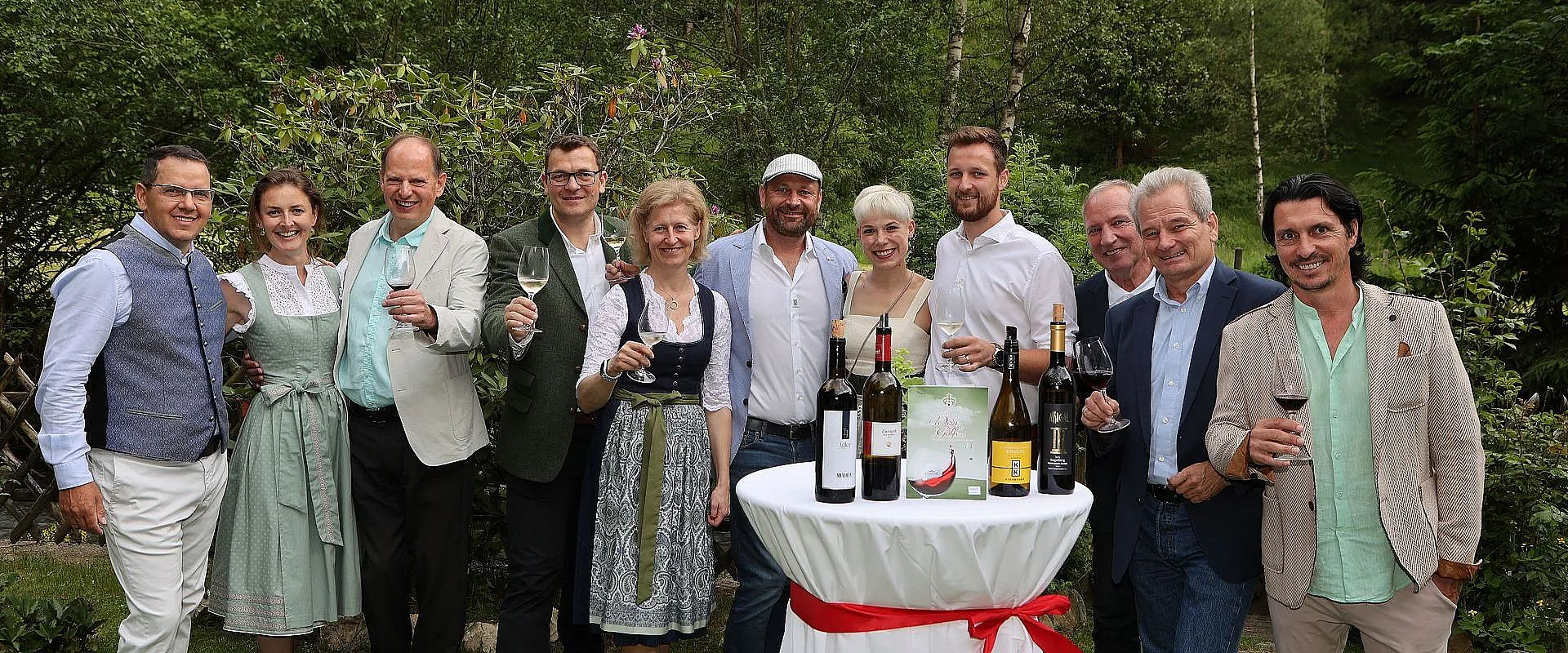 Eine Gruppe von Menschen steht fröhlich im Freien zusammen, einige halten Weingläser. Im Vordergrund ein Tisch mit mehreren Flaschen Wein darauf.