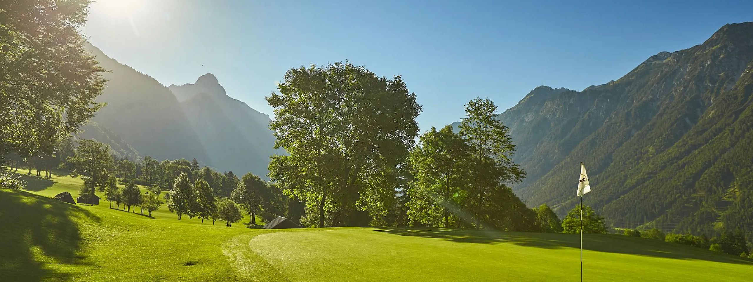 Ein gepflegtes Golfgrün mit Fahne im Sonnenlicht, umgeben von üppigen Bäumen und majestätischen Bergen im Hintergrund, unter einem klaren, blauen Himmel.