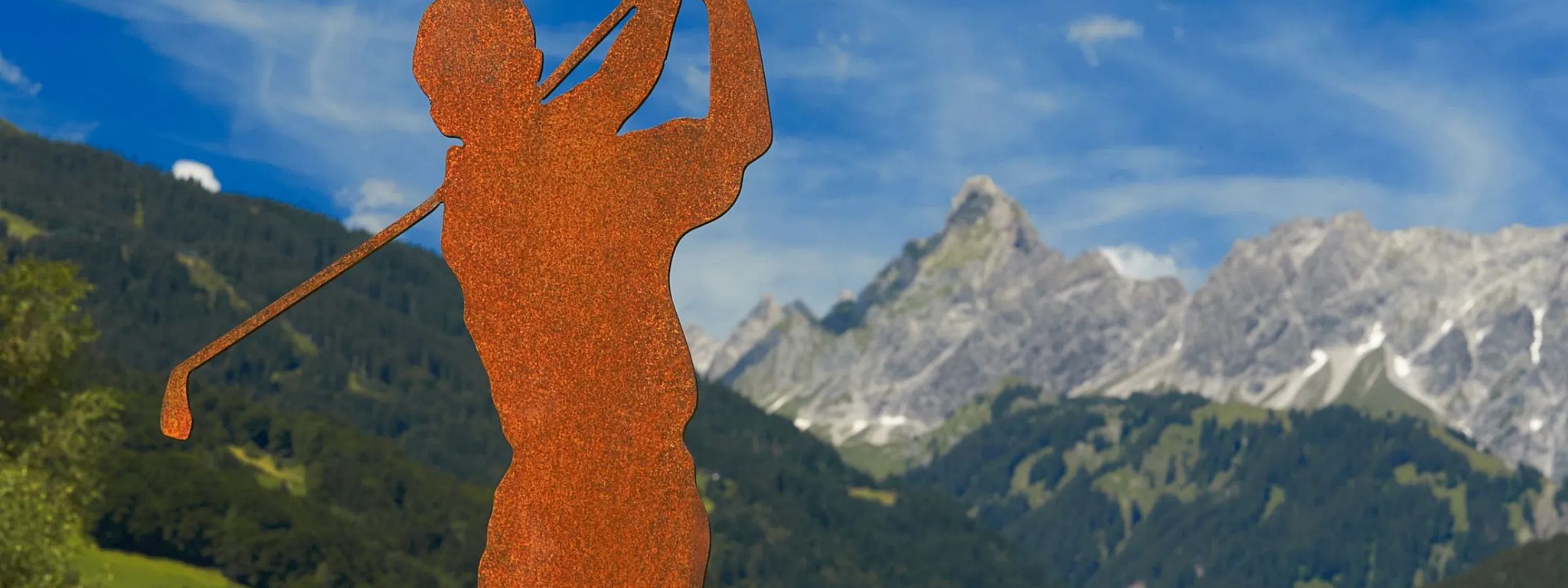 Rostige Statue eines Golfspielers vor einer beeindruckenden Berglandschaft mit grünen Hügeln und blauen Himmel im Hintergrund.