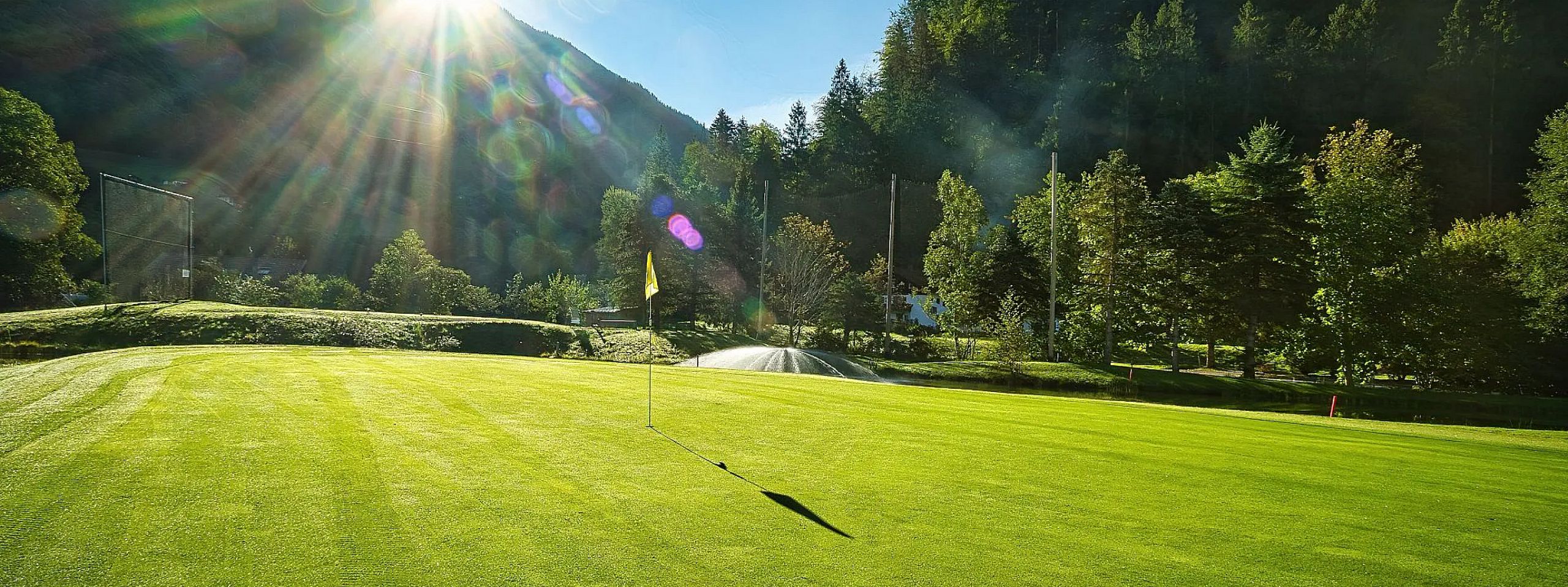 Ein sonnenbeschienener Golfplatz mit einer Fahne in der Mitte. Die Umgebung ist von dichtem Wald und Bergen umgeben, und Sonnenstrahlen durchdringen das Bild.
