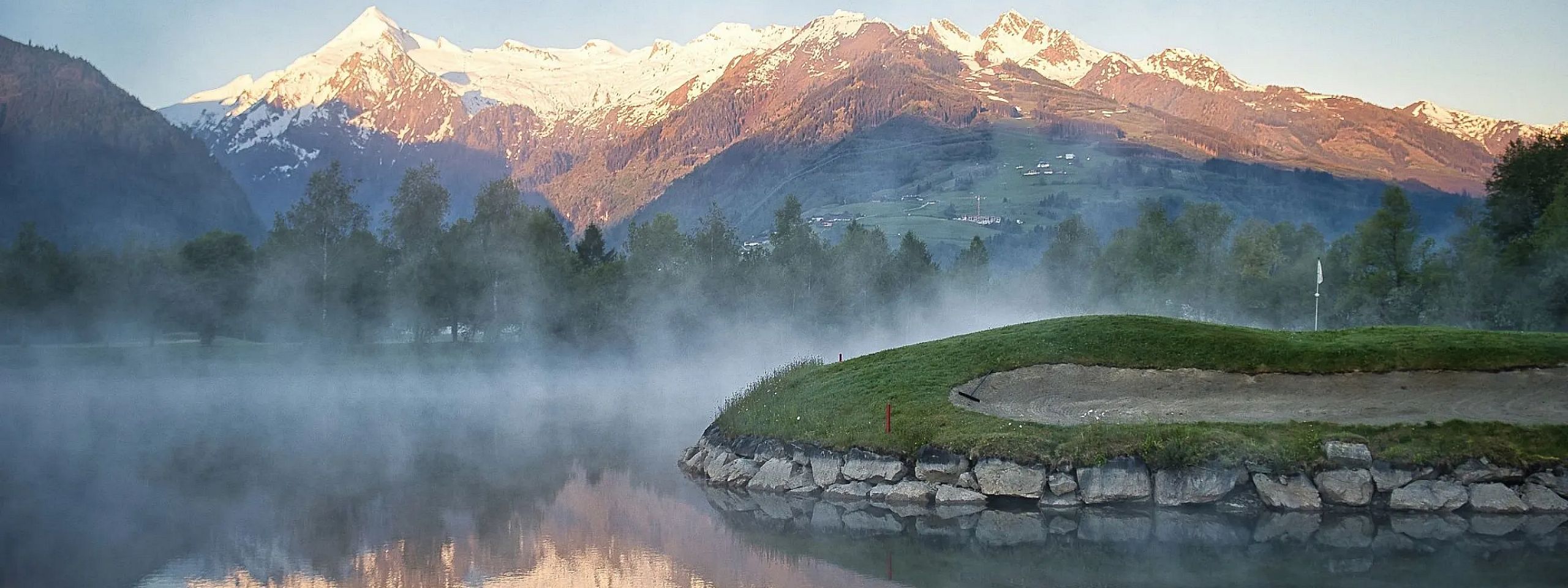 Malerische Berglandschaft mit schneebedeckten Gipfeln im Hintergrund. Im Vordergrund ein ruhiger See mit dichter Nebelbank, grasbewachsener Uferstreifen.