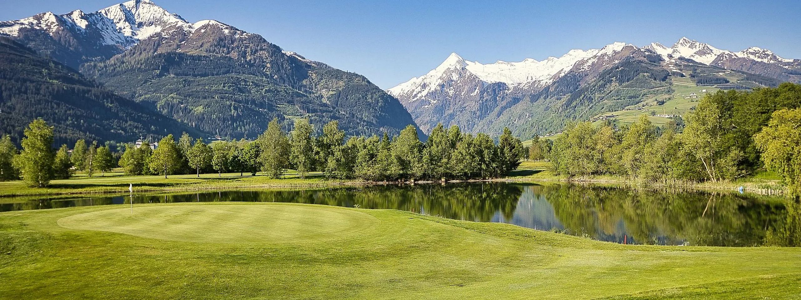 Malere Landschaft mit schneebedeckten Bergen im Hintergrund, grünem Golfplatz und ruhigem See. Der Himmel ist klar und blau, umgeben von dichter Vegetation.