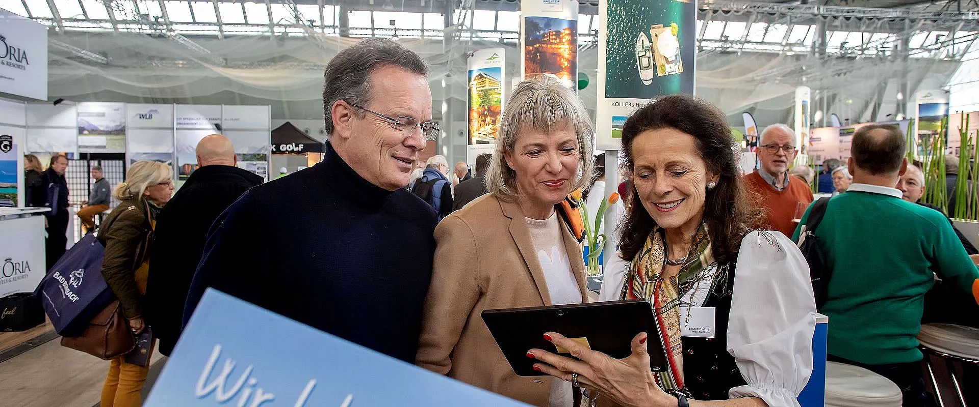 Drei Personen stehen beisammen auf einer Messe. Eine Frau zeigt den anderen beiden etwas auf einem Tablet. Im Hintergrund sind verschiedene Messestände zu sehen.