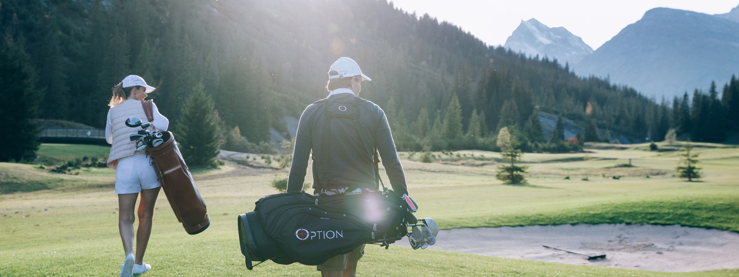 Golfer in sportlicher Kleidung trägt seine Golftasche über der Schulter, während er über einen grünen Golfplatz im Gebirge geht. Die Sonne scheint leicht durch die Bäume.