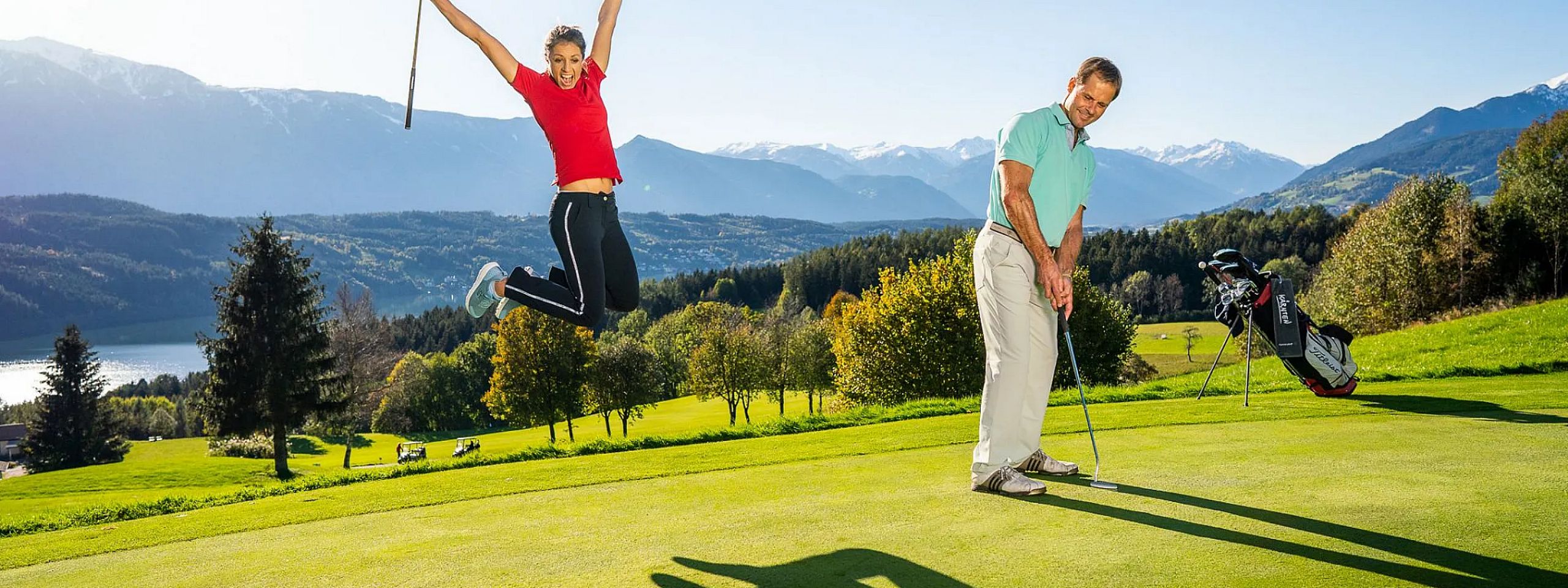 Zwei Personen auf einem Golfplatz: Eine Frau in Rot springt freudig in die Luft, während ein Mann konzentriert puttet. Hintergrund mit Bergen und Bäumen.