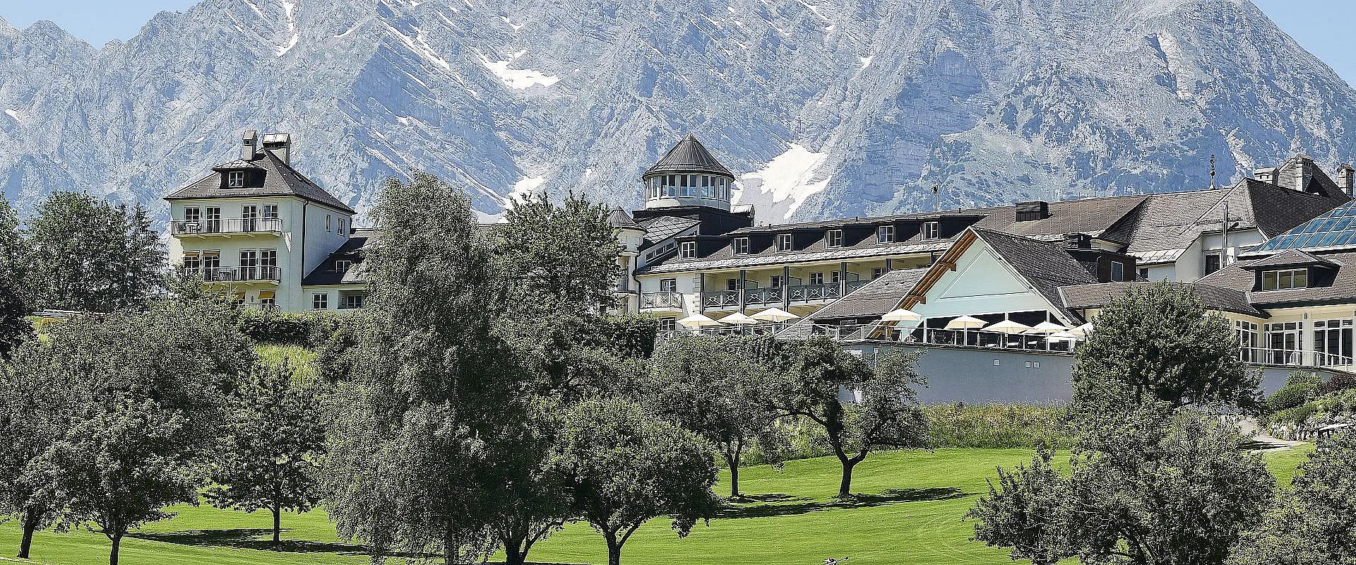 Ein elegantes Hotel mit Satteldach steht vor einer beeindruckenden Bergkulisse. Üppige grüne Bäume und Wiesen umgeben das Gebäude an einem klaren Tag.