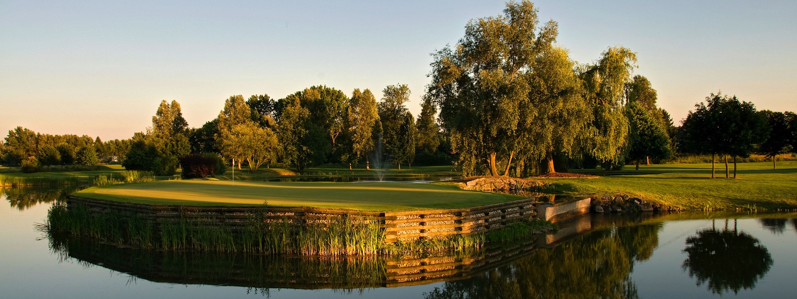 Eine idyllische Golfplatzlandschaft bei Sonnenuntergang, umgeben von Bäumen und einem ruhig spiegelnden See, der die friedliche Atmosphäre verstärkt.