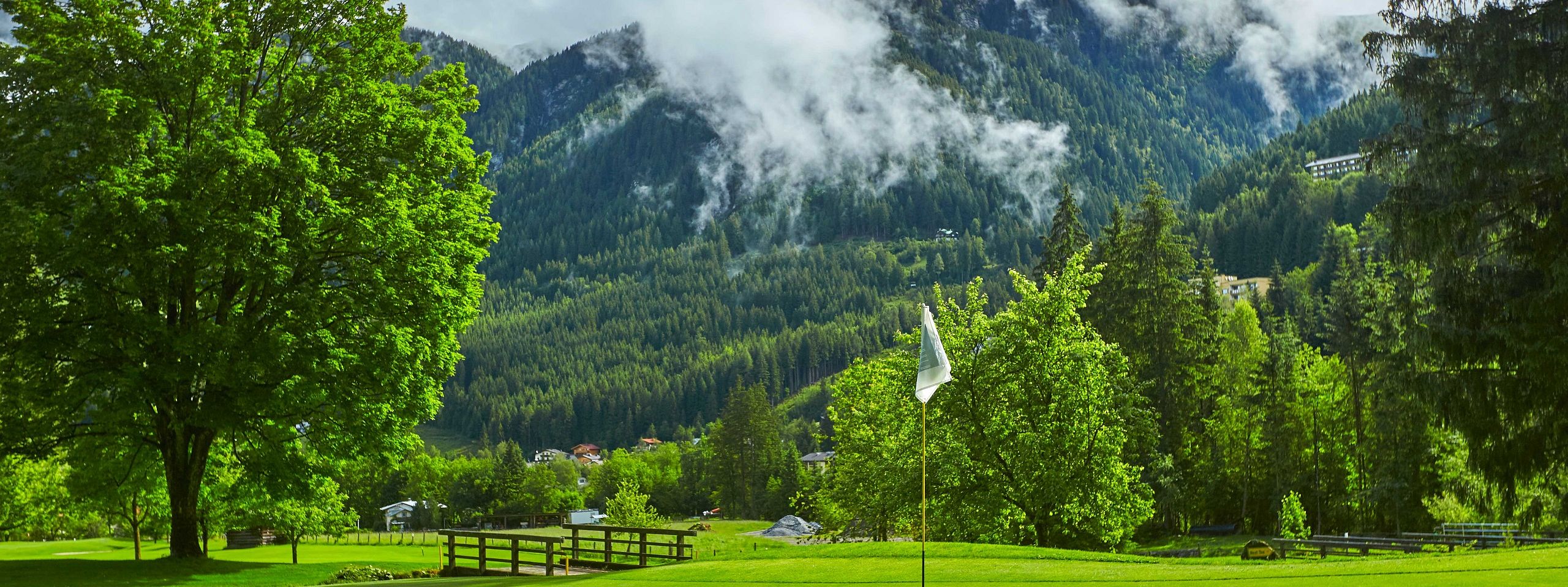 Lush golf course with a flag in the foreground, surrounded by trees and mountains under a partly cloudy sky.