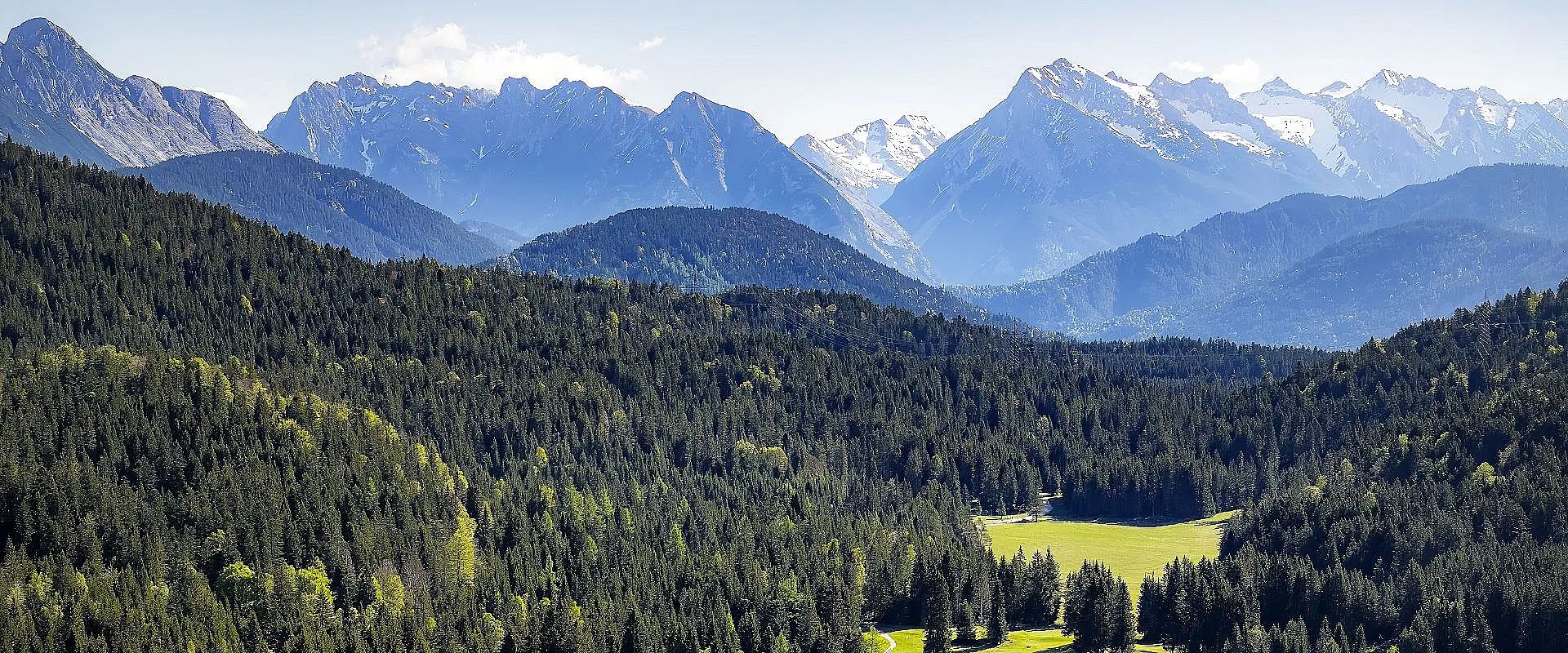 Ein malerisches Bergpanorama mit verschneiten Gipfeln im Hintergrund, dichten Wäldern und grünen Wiesen im Vordergrund unter einem blau bewölkten Himmel.
