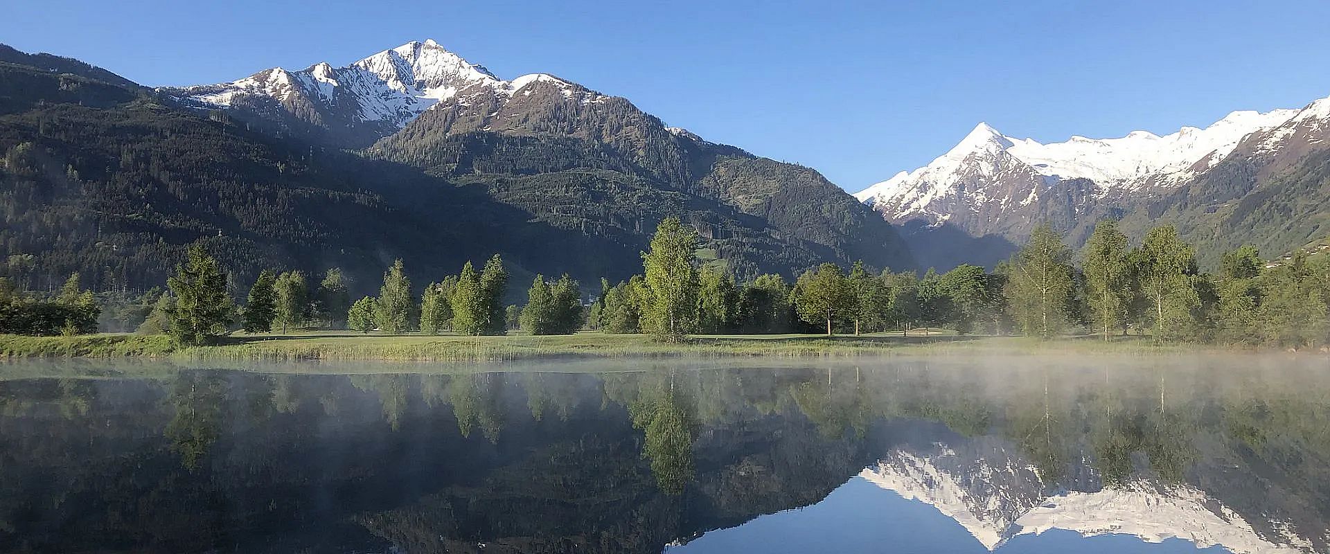Ein klarer Bergsee spiegelt schneebedeckte Alpengipfel und grüne Wälder wider, während Nebel sanft über der Wasseroberfläche schwebt. Blauer Himmel im Hintergrund.